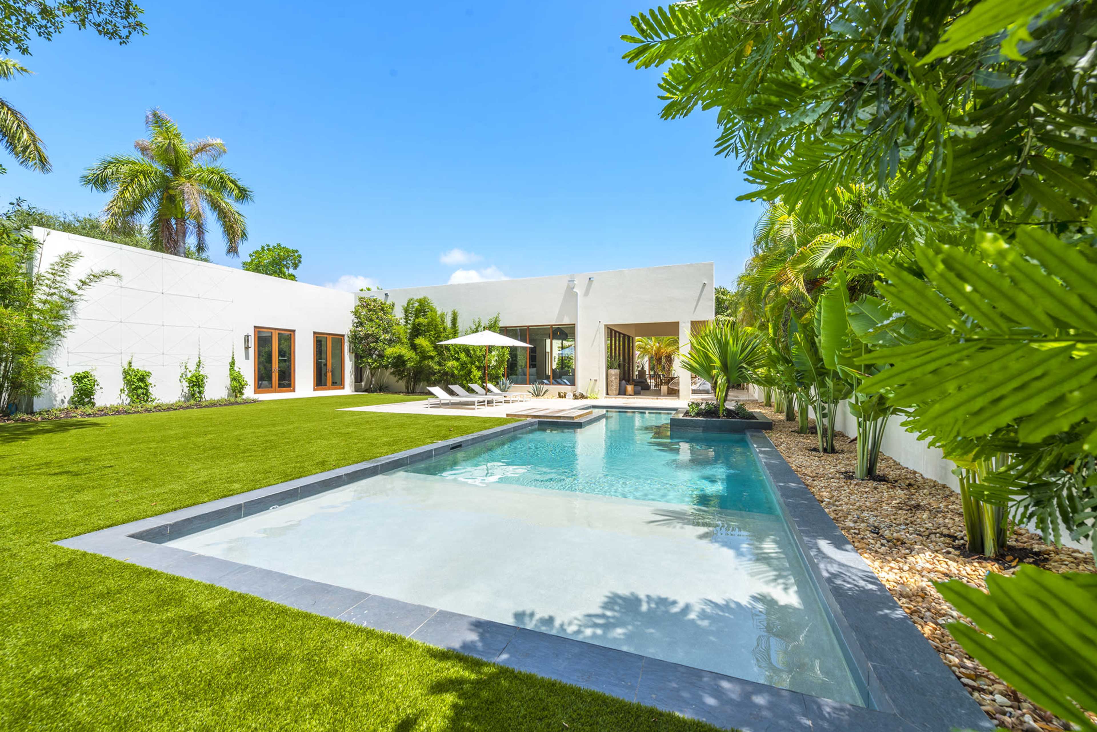 A modern house with large windows overlooks a swimming pool surrounded by a well-manicured lawn and tropical plants.