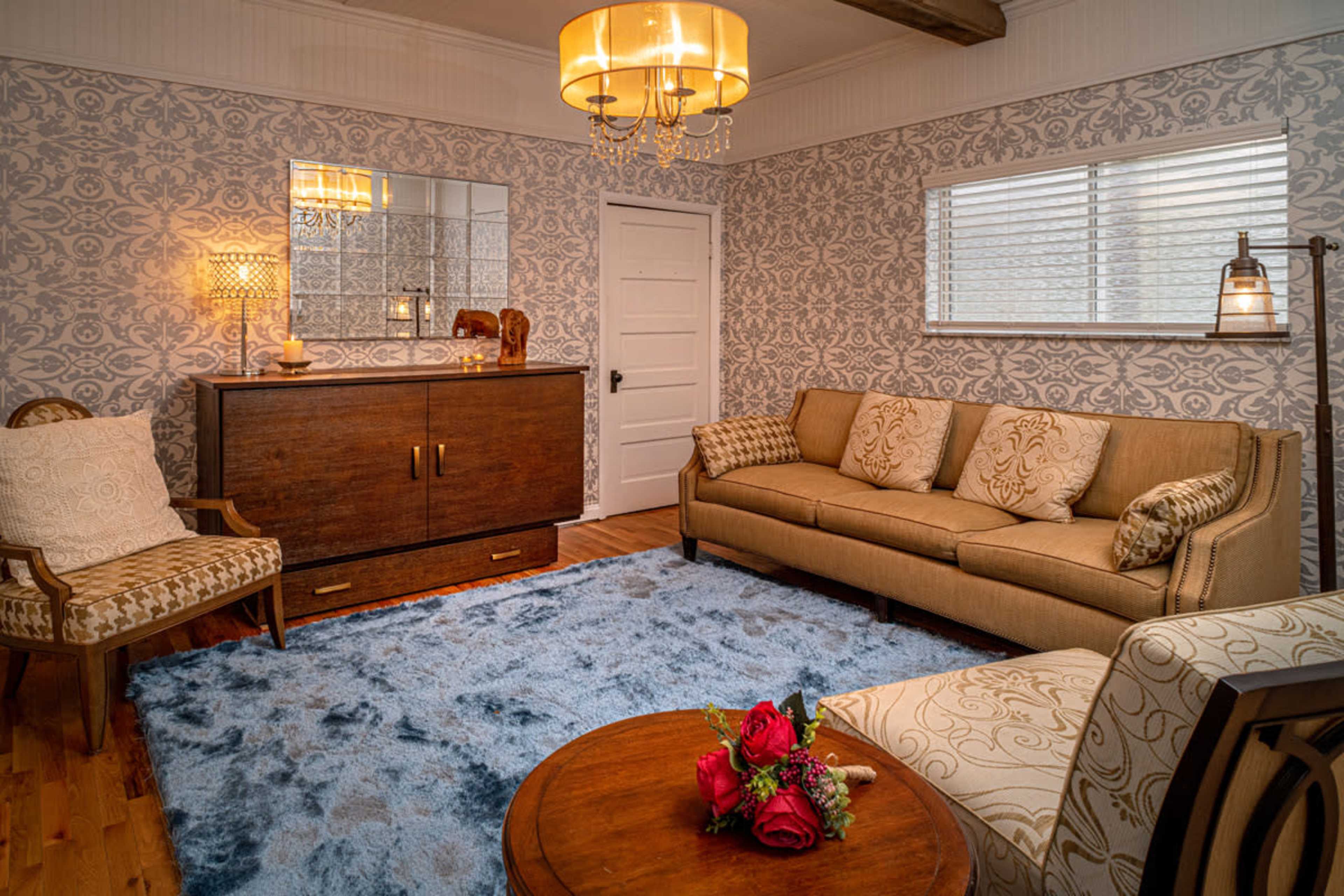 The room features a light blue rug, a beige sofa adorned with patterned cushions, a wooden coffee table with flowers, and a decorative chest against a patterned wall.