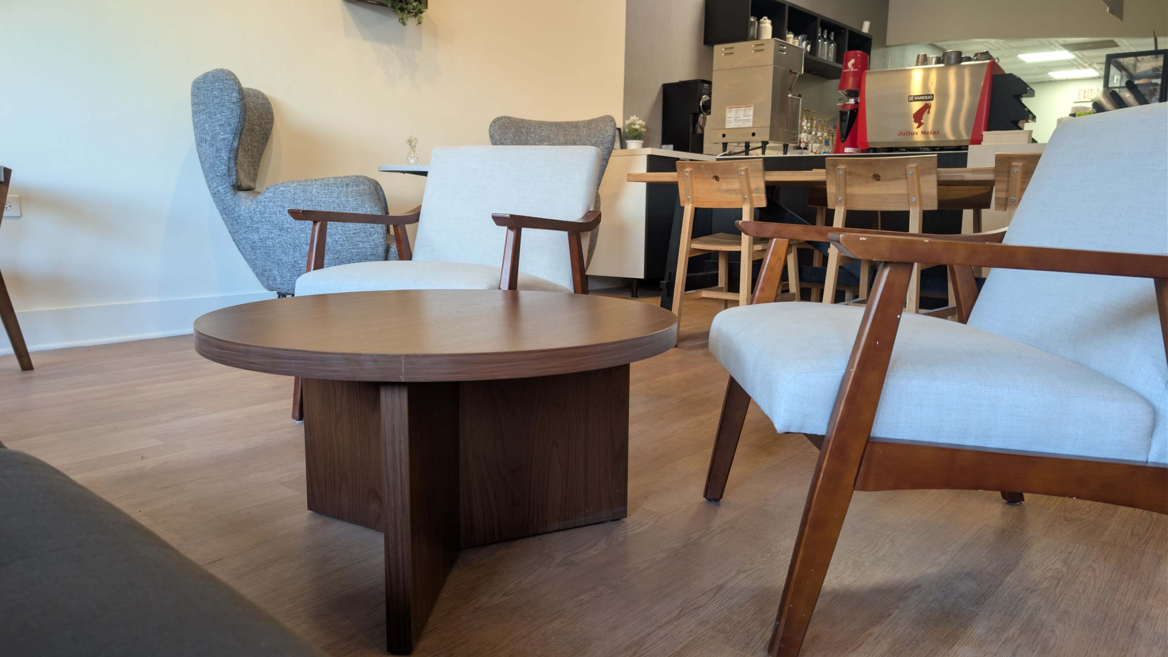 The image shows a cozy seating area with two armchairs and a round wooden coffee table, set against a background of a kitchen and dining space.