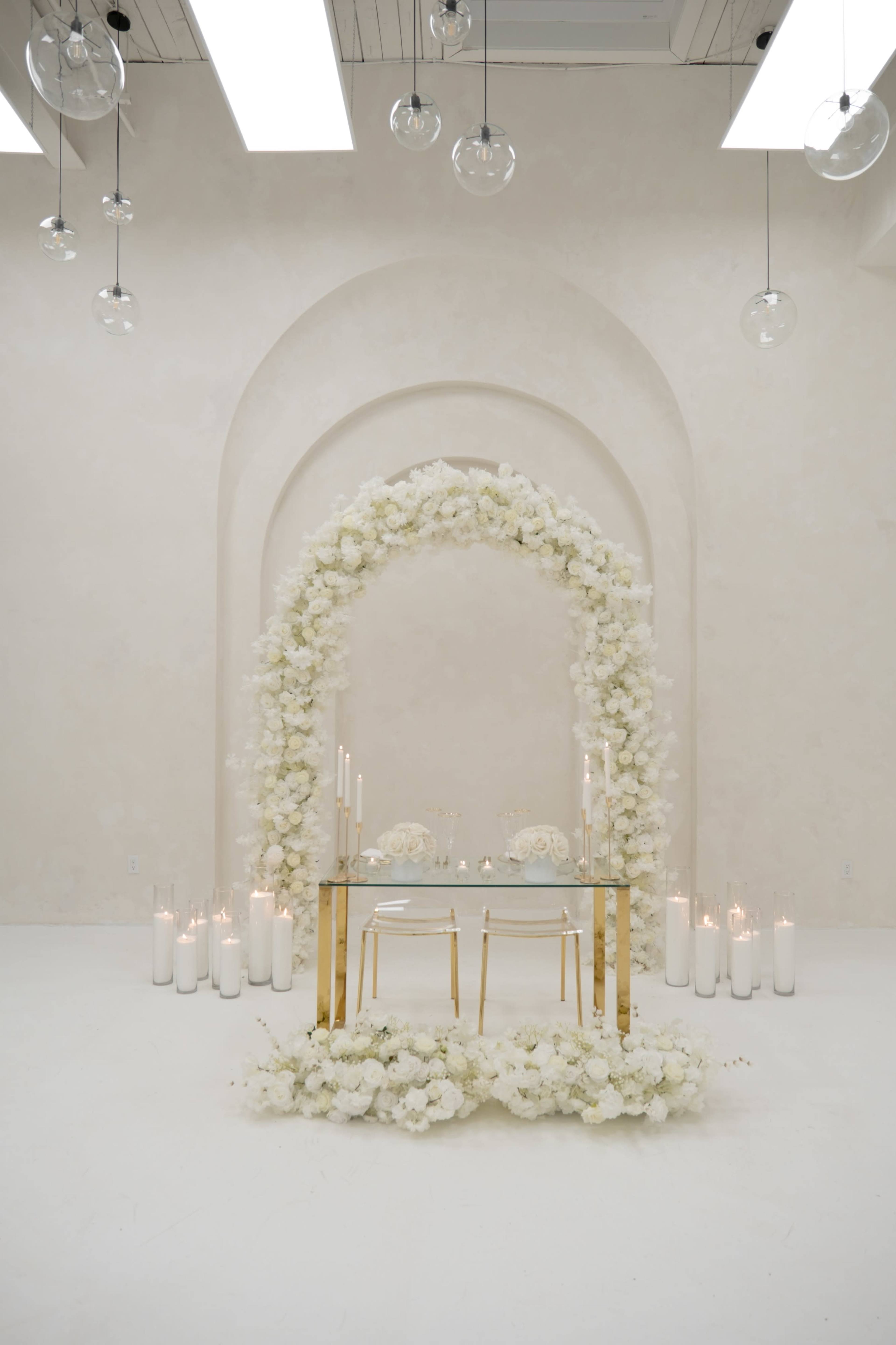 A minimalist wedding altar features a floral archway adorned with white flowers, surrounded by candles on the floor in a softly lit, neutral-toned room.