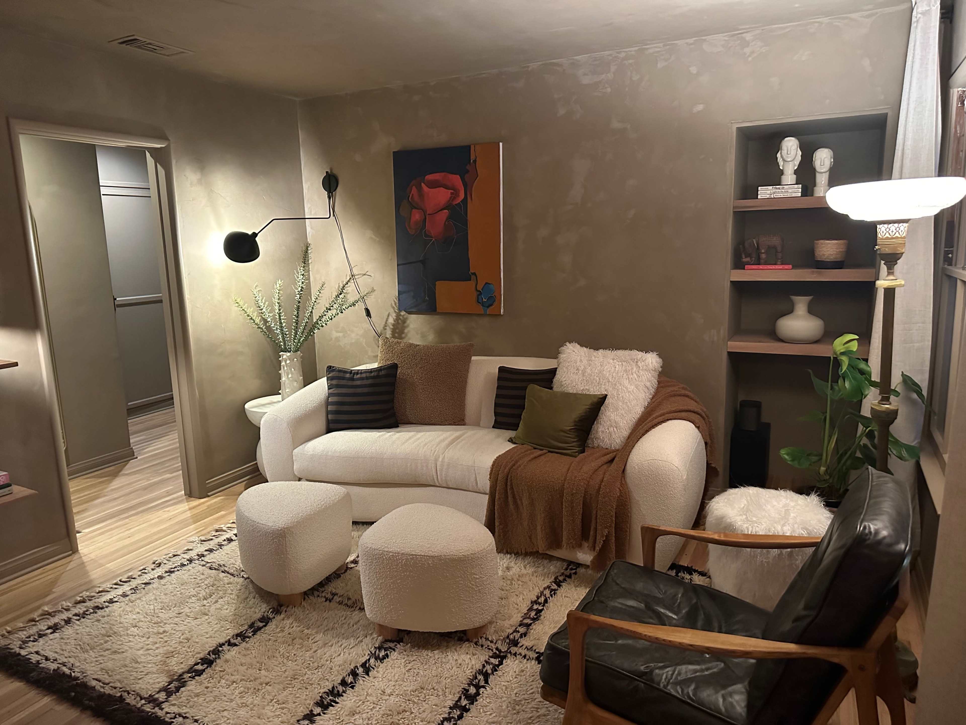 The image shows a cozy living room featuring a white sofa with decorative pillows, two round ottomans, a green armchair, and a wall-mounted lamp, complemented by wall art and plants.