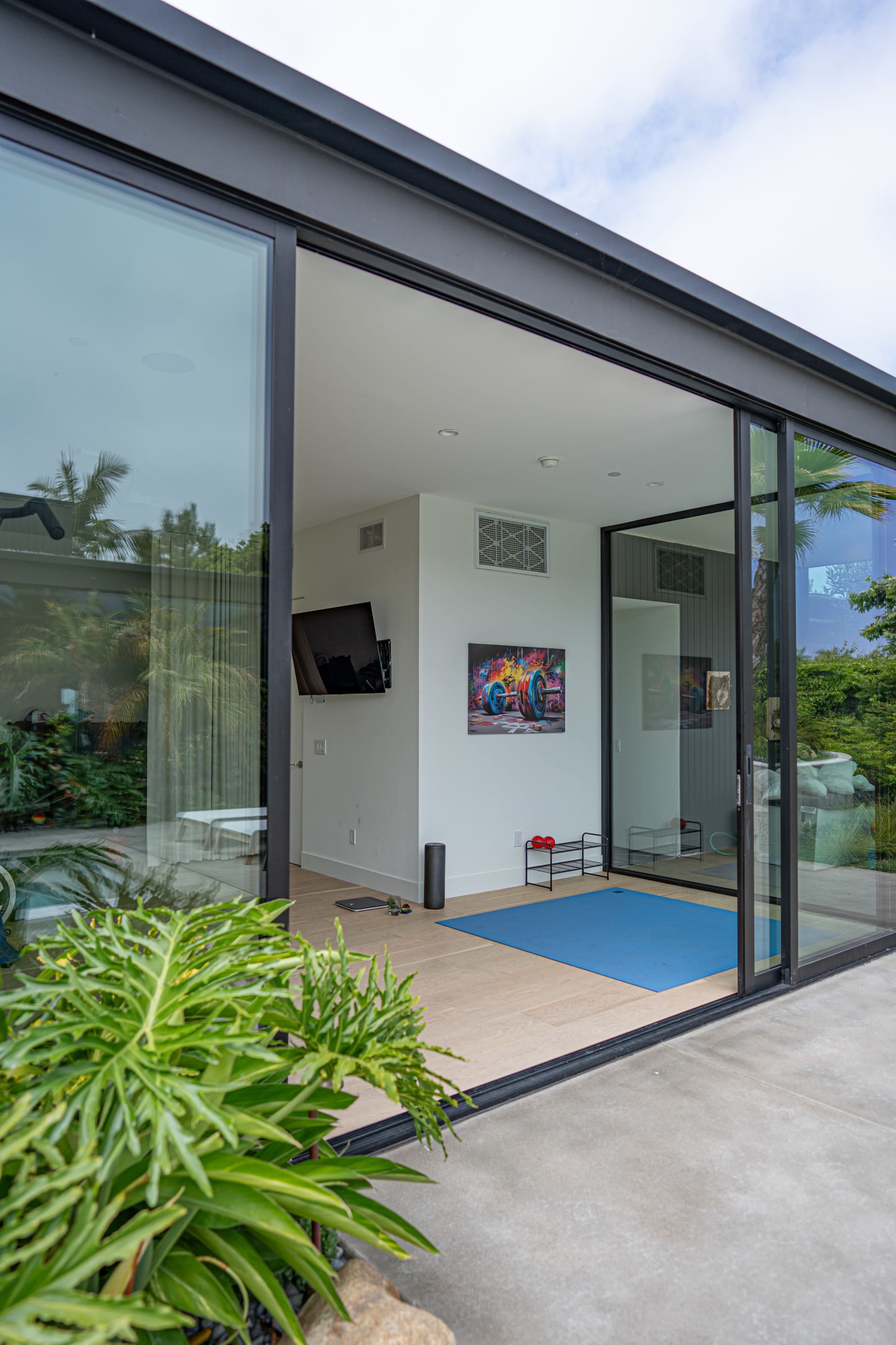 The image shows a modern home with large glass walls revealing a gym area featuring exercise equipment and artwork.