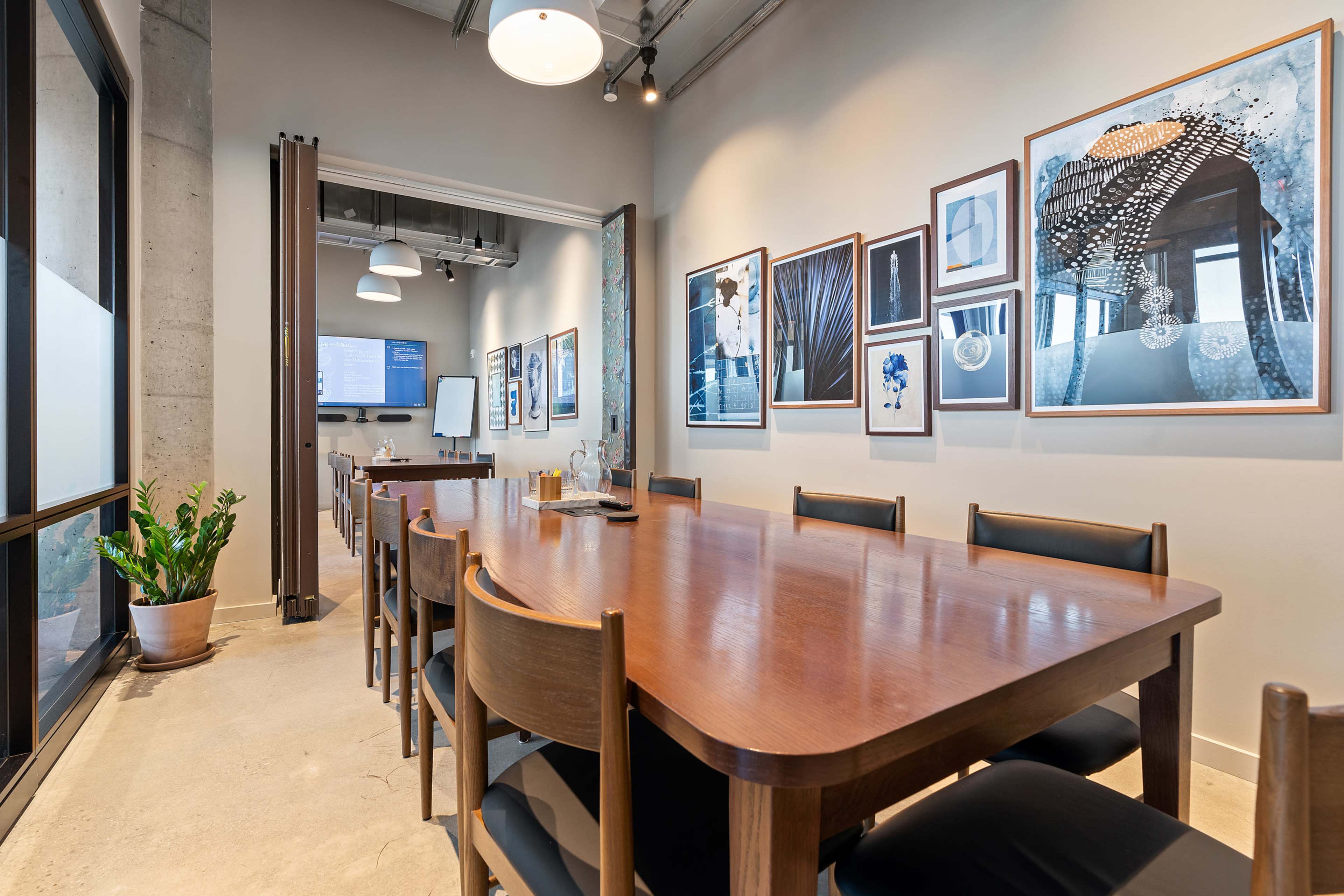 A long wooden conference table sits in a well-lit meeting room adorned with framed artwork on the walls.
