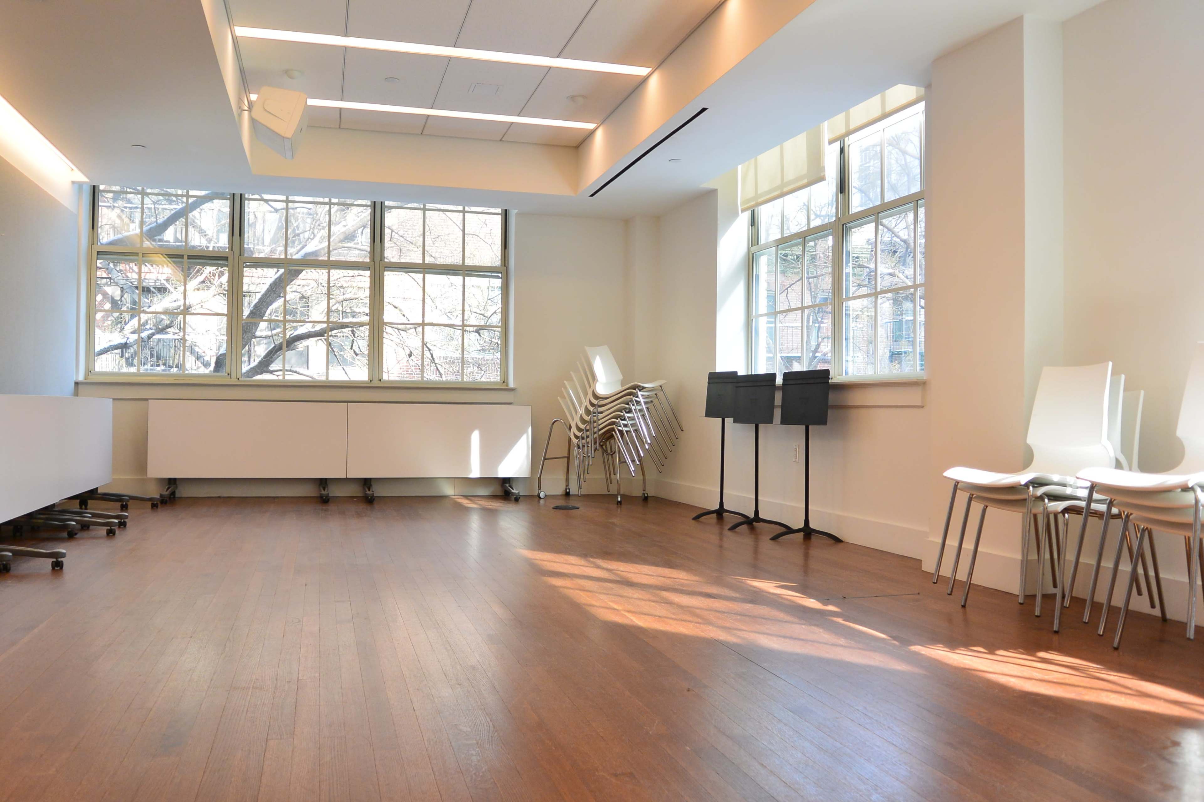 The image shows a bright, empty room with wooden floors, large windows, and neatly stacked white chairs against one wall.