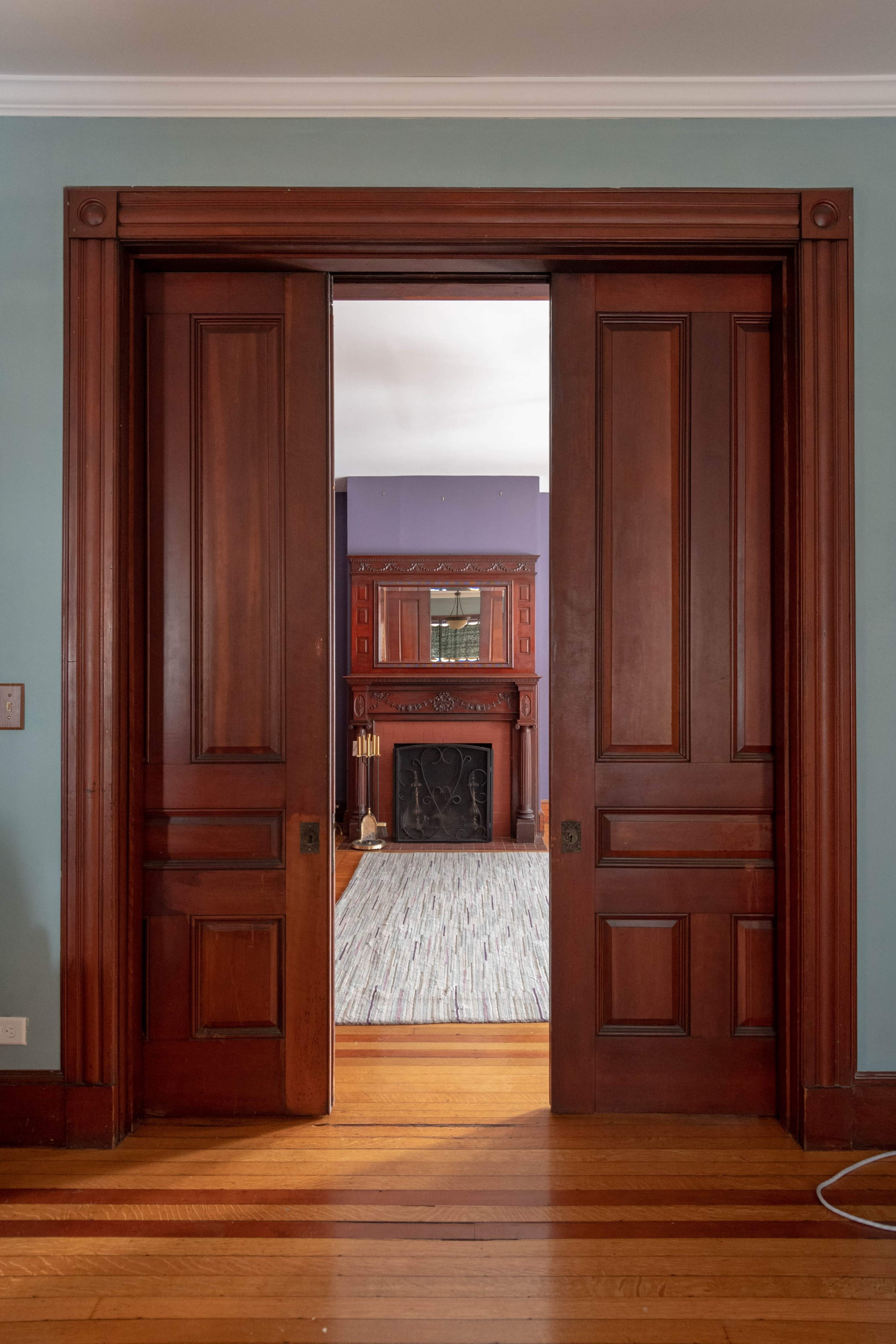 A pair of wooden doors opens to reveal a room featuring a vintage fireplace and patterned carpet.