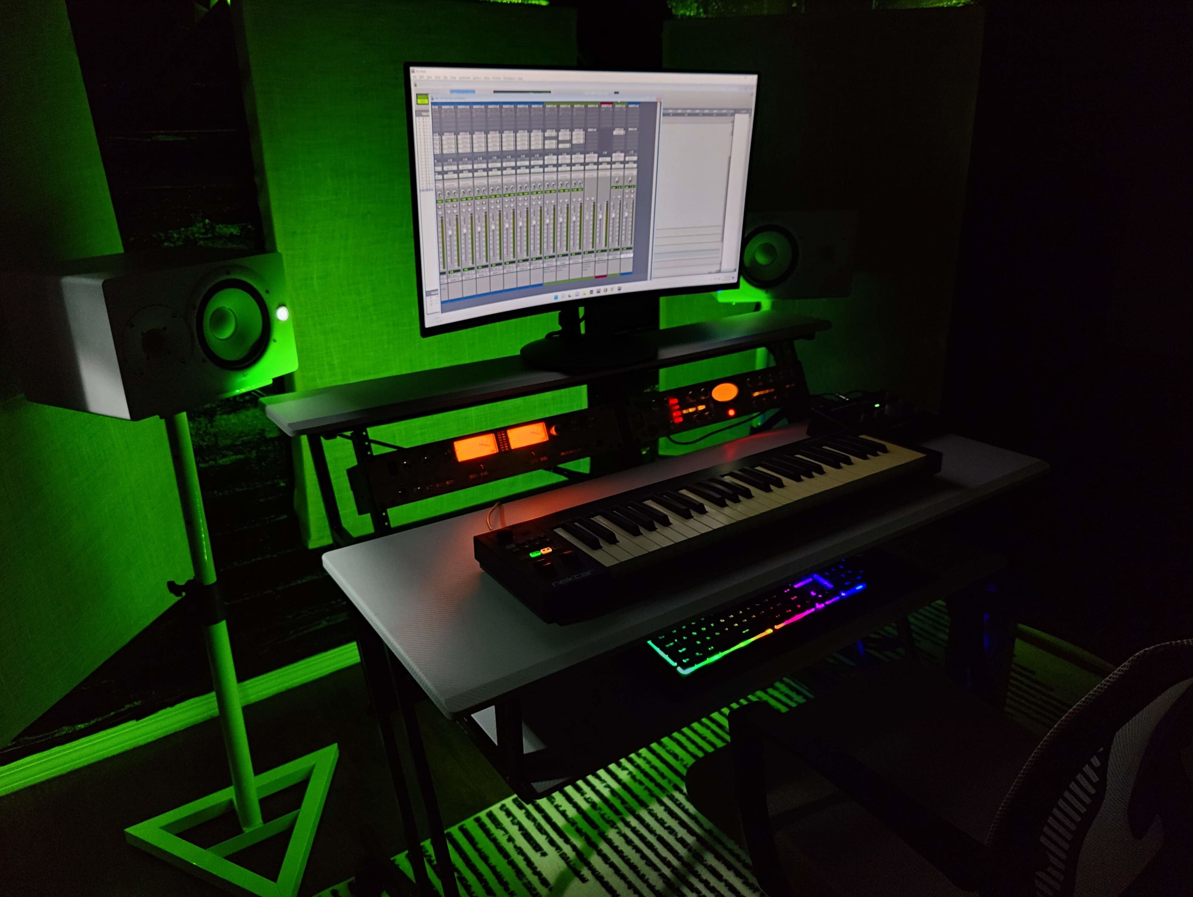 A home studio setup features a keyboard, computer monitor displaying audio software, and speakers, all illuminated with green lighting.