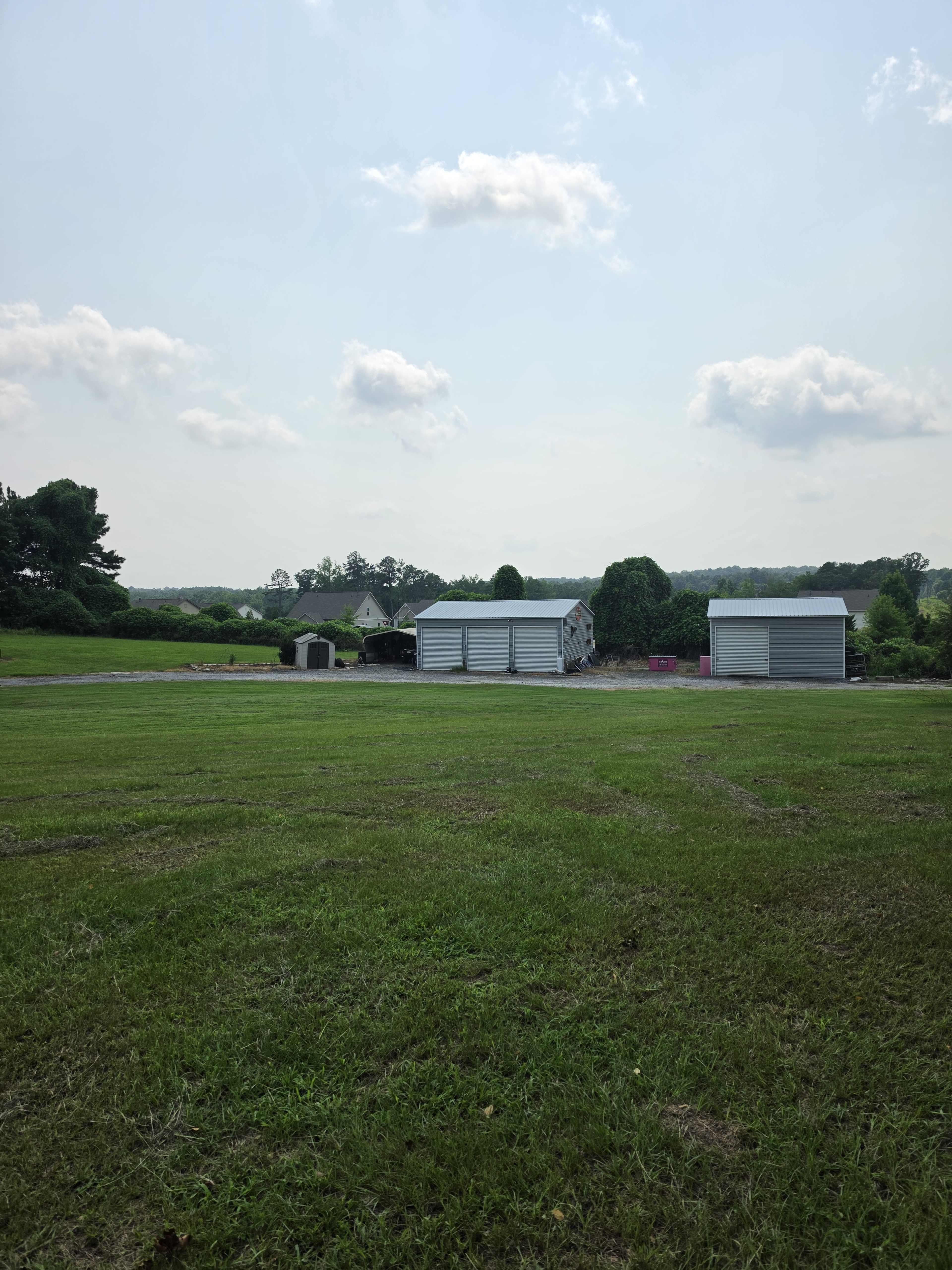 The image shows a grassy field with two large, gray storage buildings in the background, set against a blue sky with a few clouds.