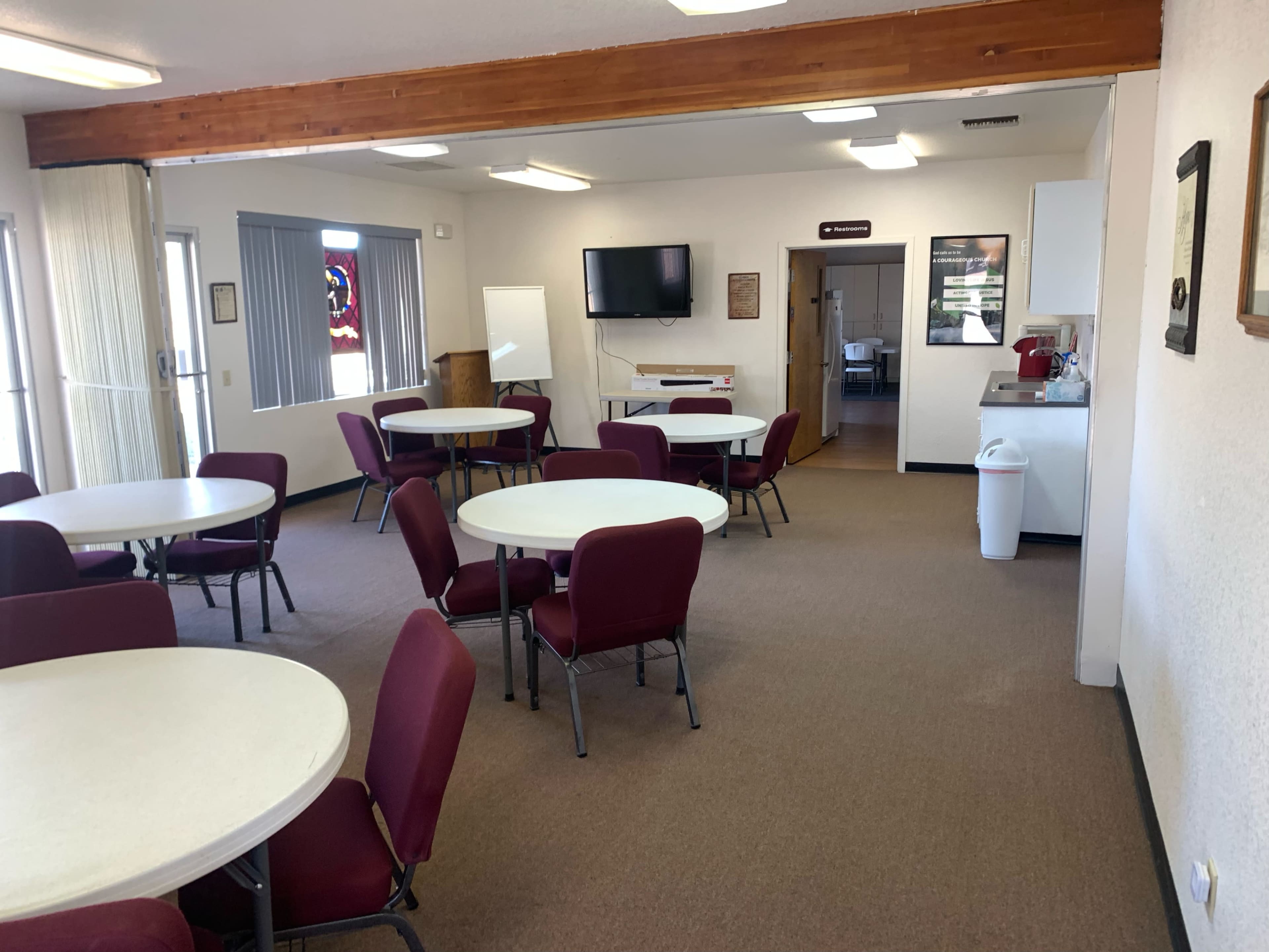 A large, well-lit room with several round tables and maroon chairs, along with a kitchenette area and a wall-mounted television.