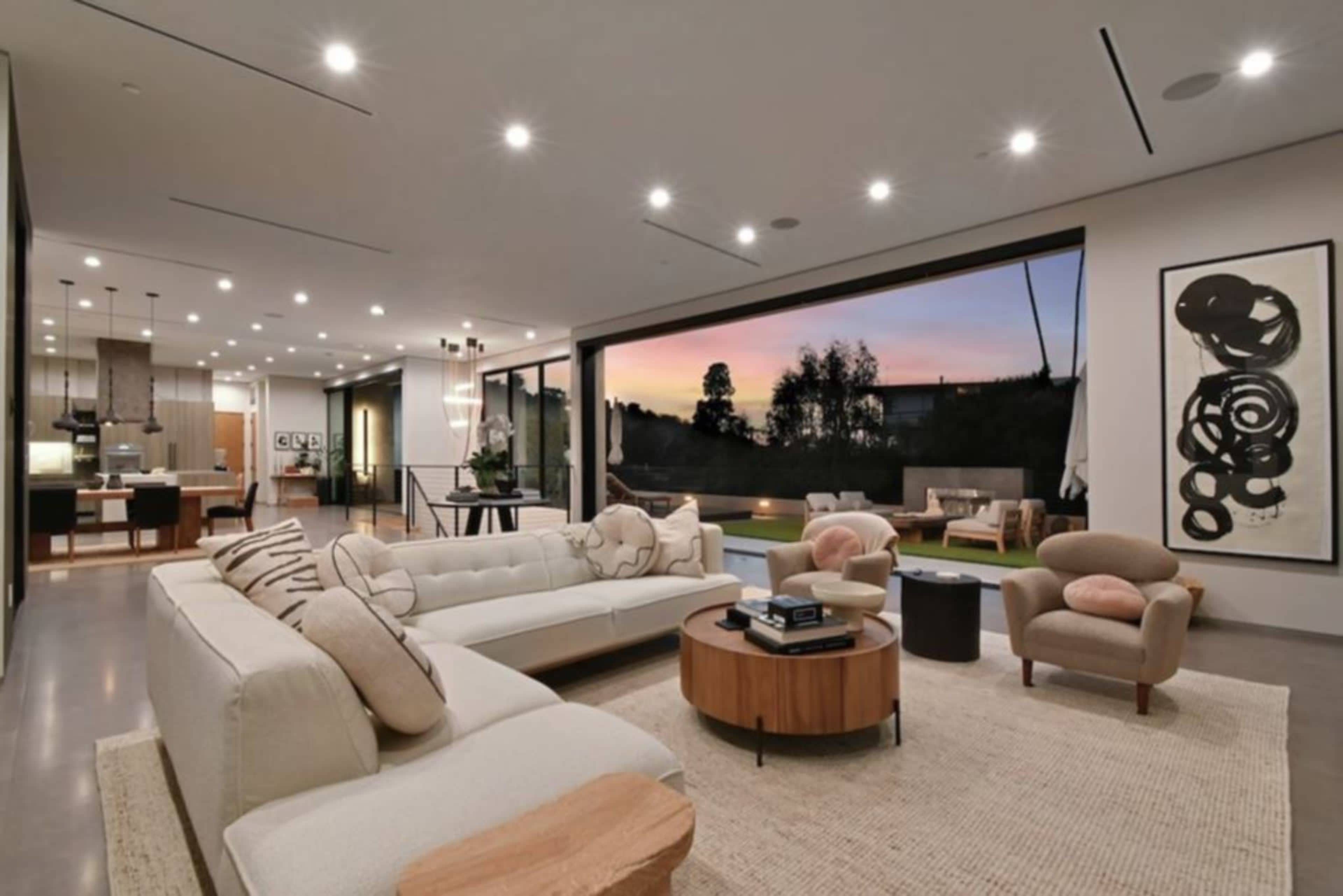 The image depicts a modern living room featuring a large sectional sofa, a round coffee table, and large sliding glass doors that open to a view of a sunset.