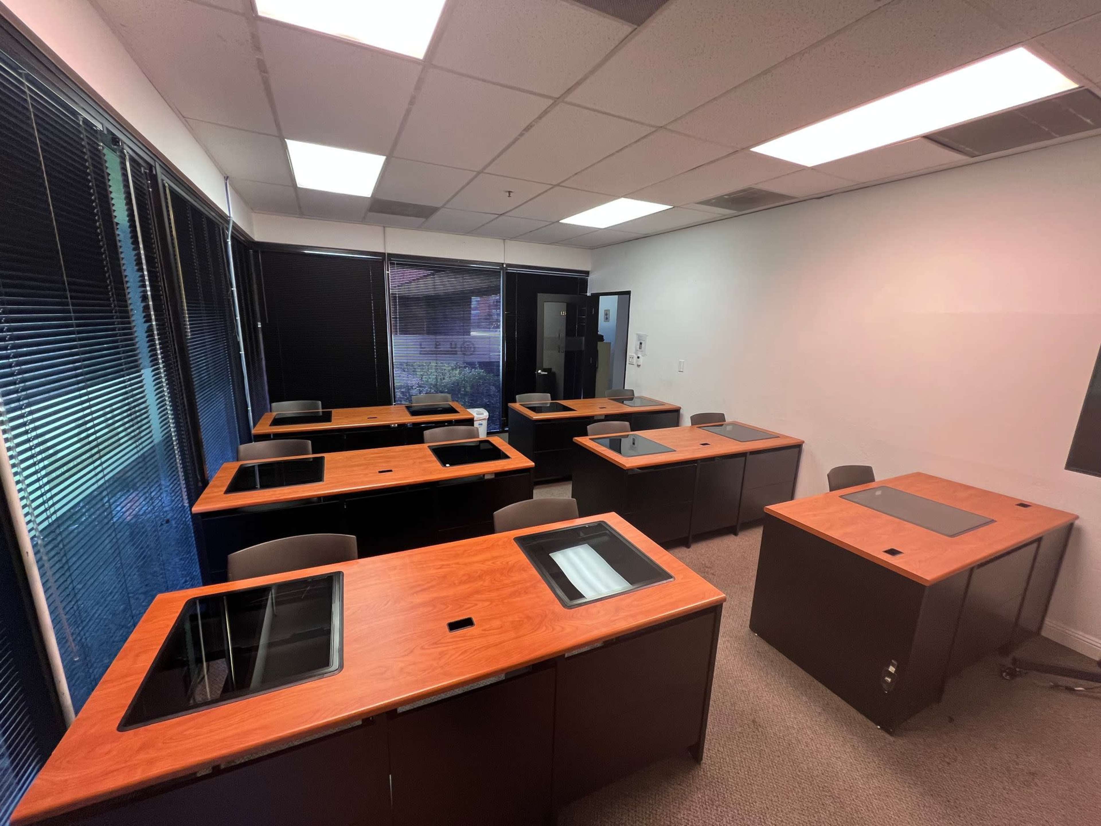 The image shows a classroom or office space with multiple wooden desks arranged in rows, each equipped with a black workspace and windows providing natural light.