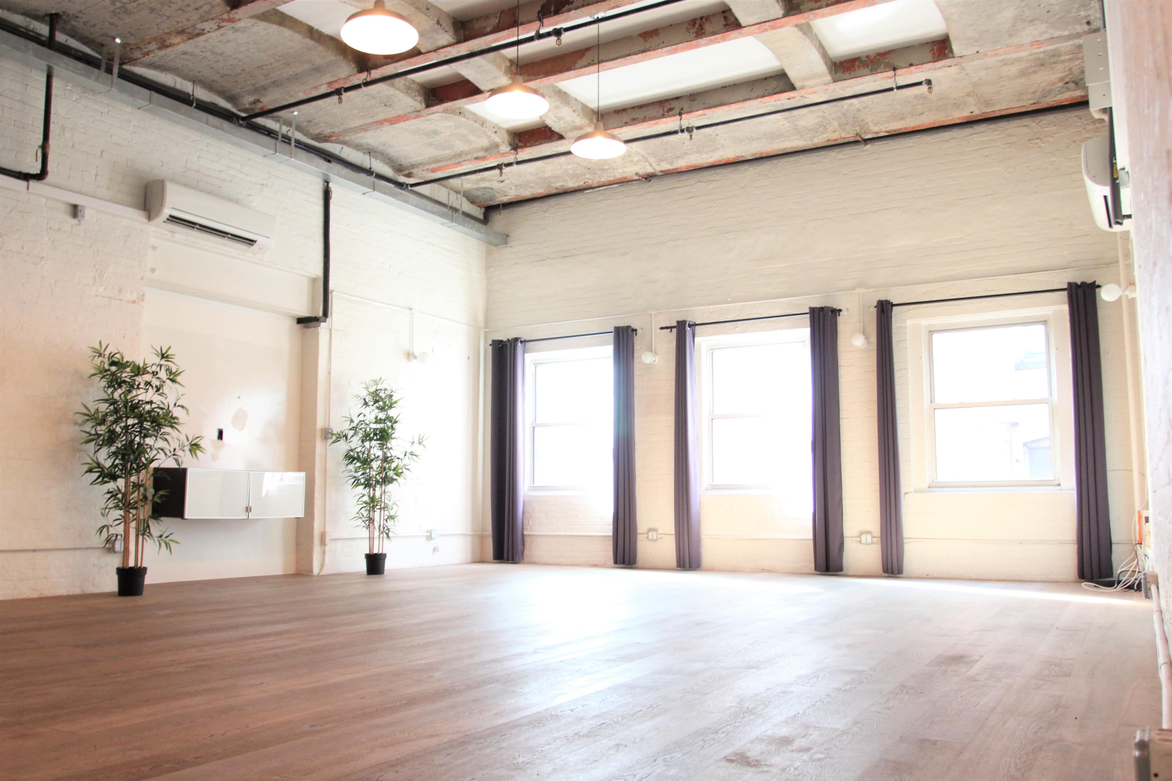 A spacious, empty room with wooden flooring, large windows, and potted plants in the corners.