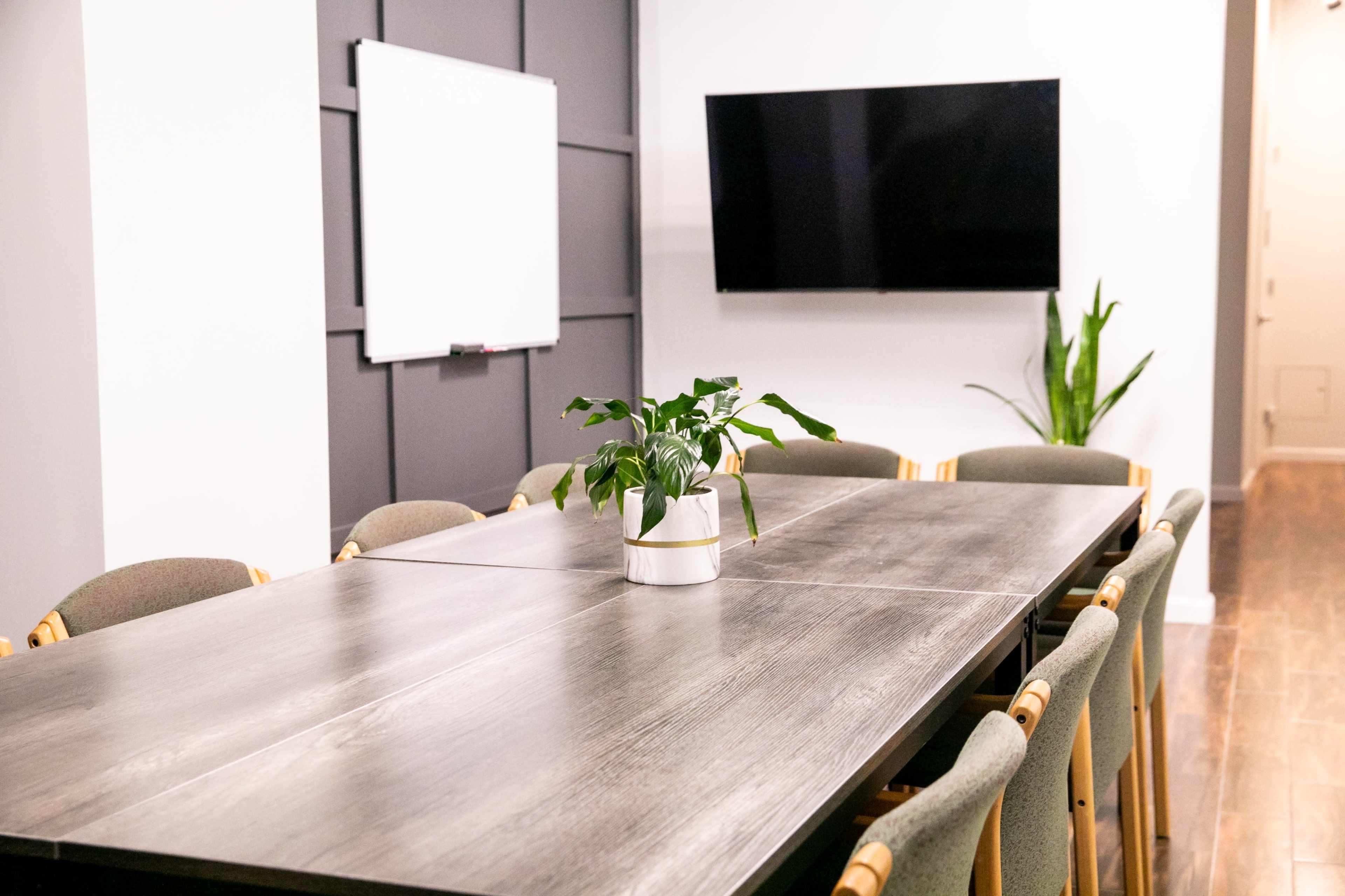 A modern conference room features a large wooden table surrounded by chairs, with a plant centerpiece, a whiteboard, and a television on the wall.