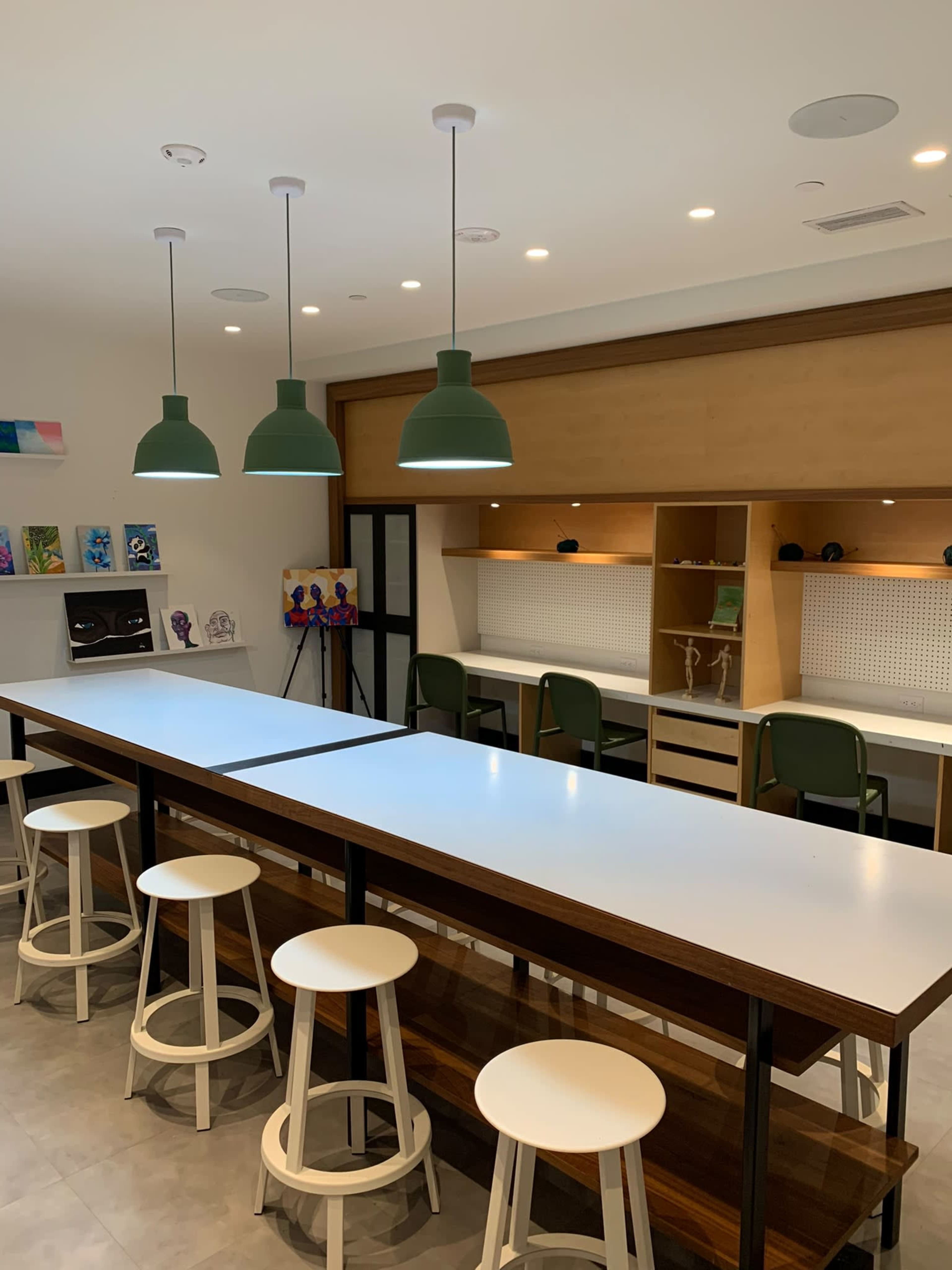 The image shows a modern kitchen with a long white countertop illuminated by green pendant lights, accompanied by white stools and wooden accents.