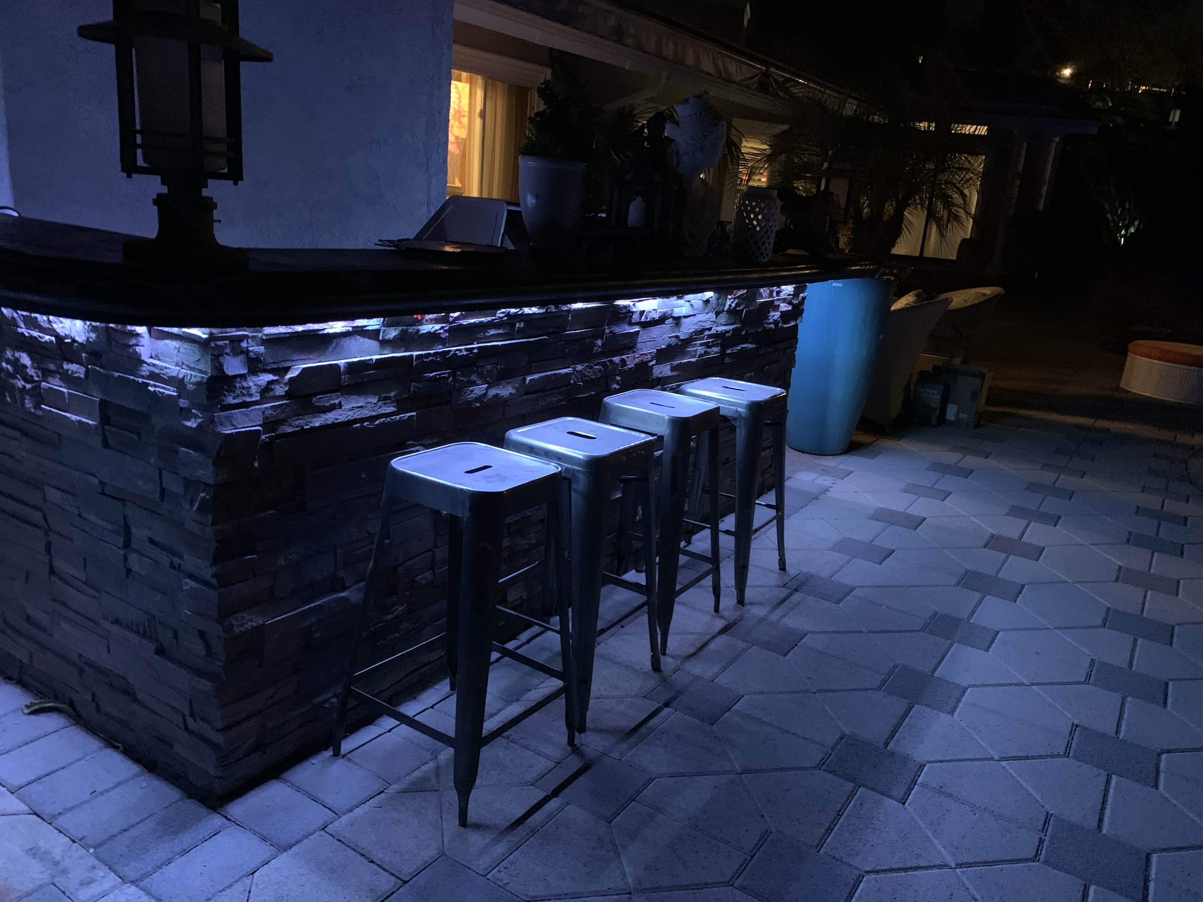 A dimly lit outdoor bar area with three metal stools placed in front of a stone bar counter.