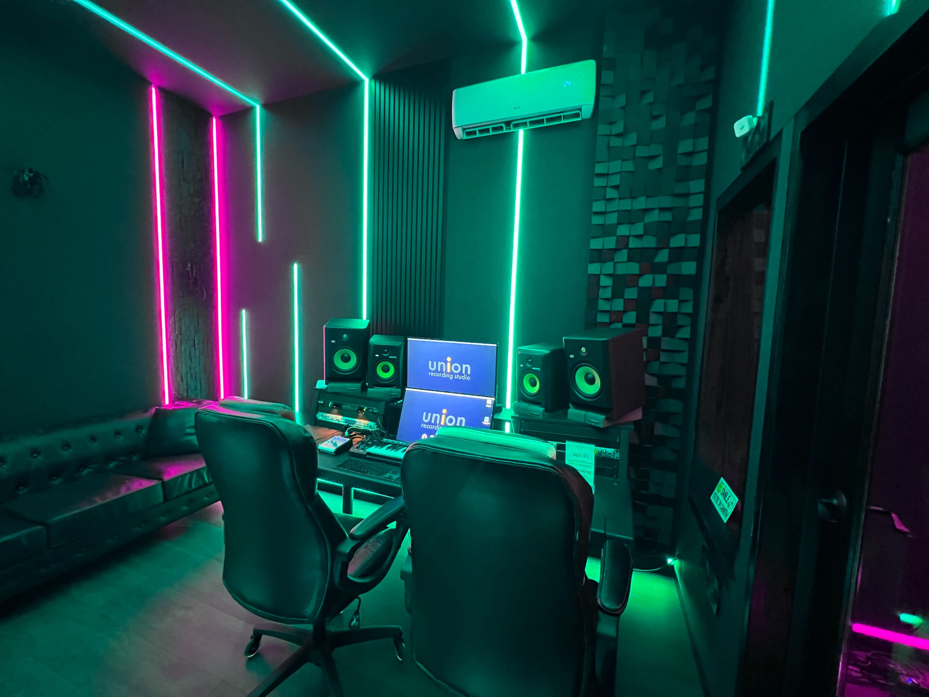 A recording studio with glowing neon lights, two black chairs, a computer setup, and audio speakers against a dark backdrop.