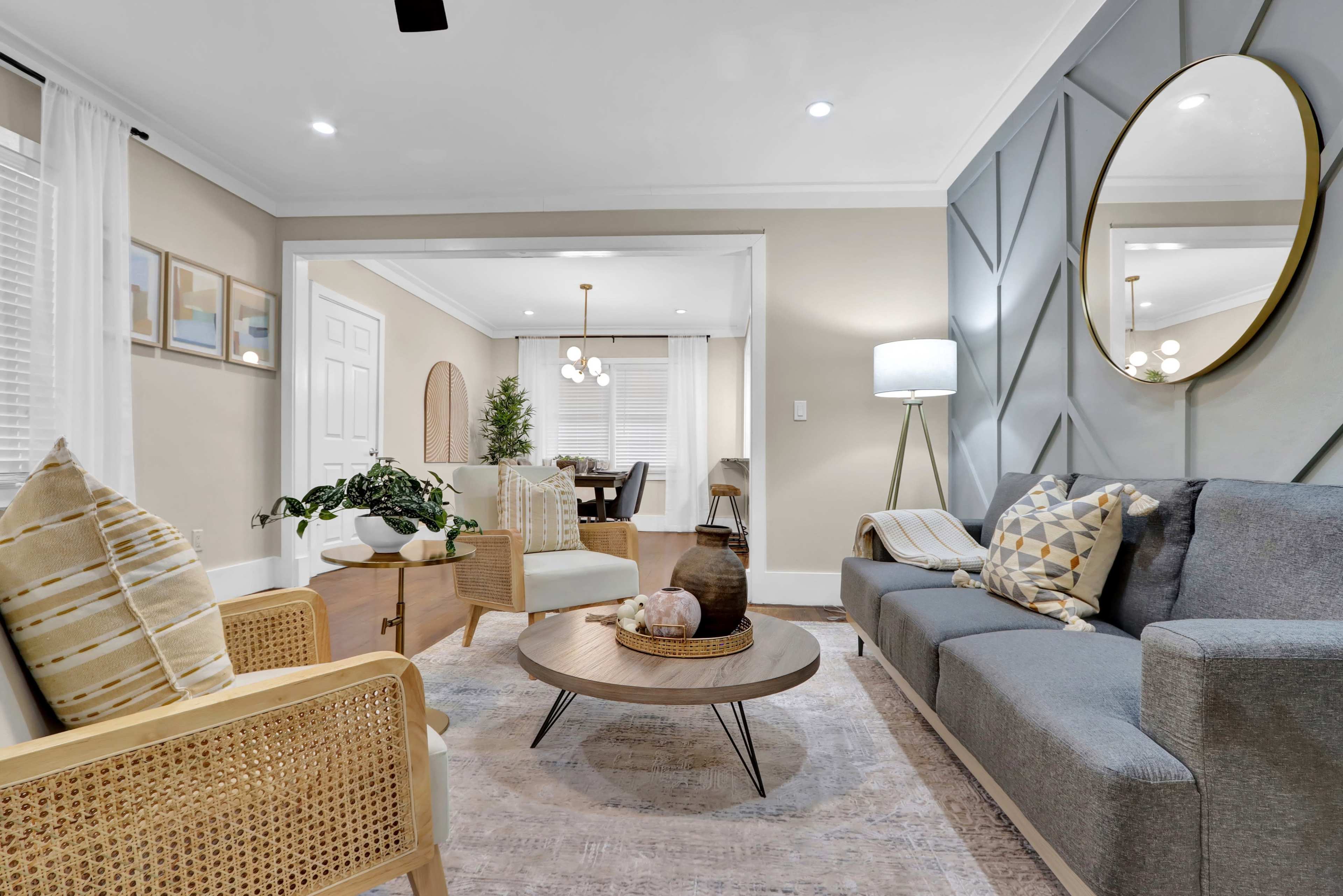 A modern living room features a gray sofa, a round coffee table, and a decorative mirror on the wall, with natural light illuminating the space through large windows.