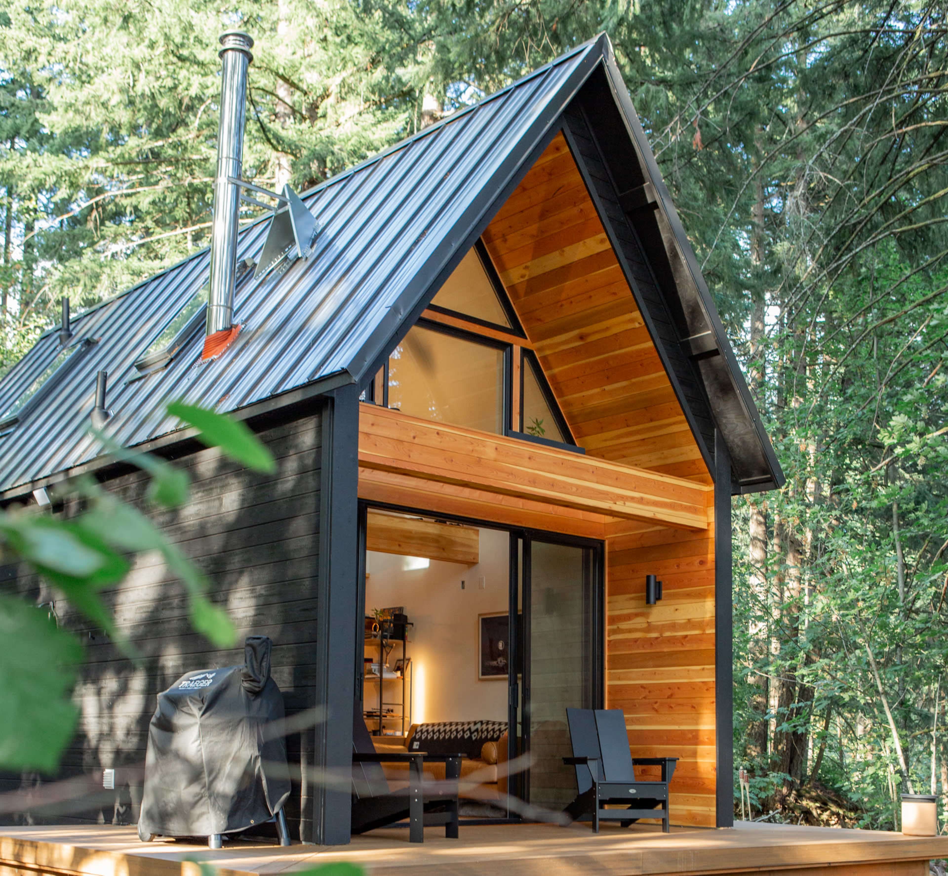 A modern cabin with a sloped metal roof and wooden accents is set amidst tall trees, featuring large glass doors that open onto a wooden deck.