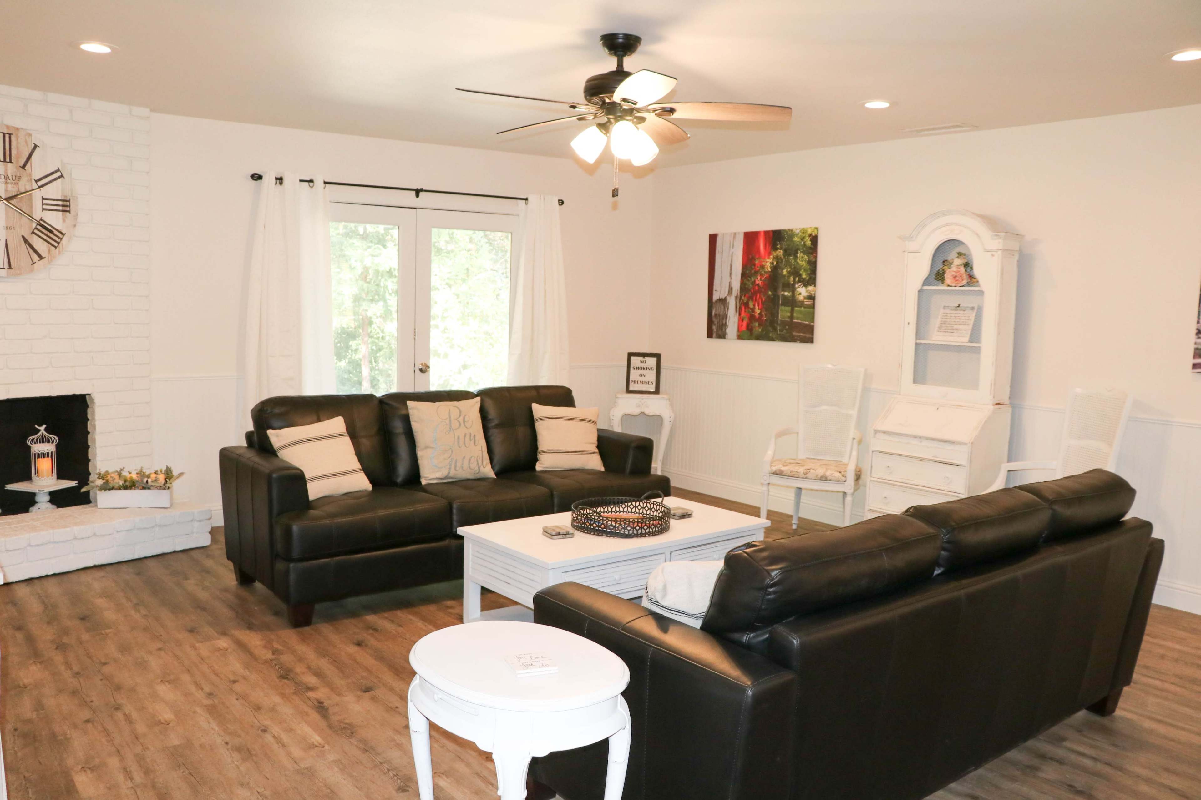A living room features two black leather sofas, a white coffee table, a white brick fireplace, and large windows with light curtains.