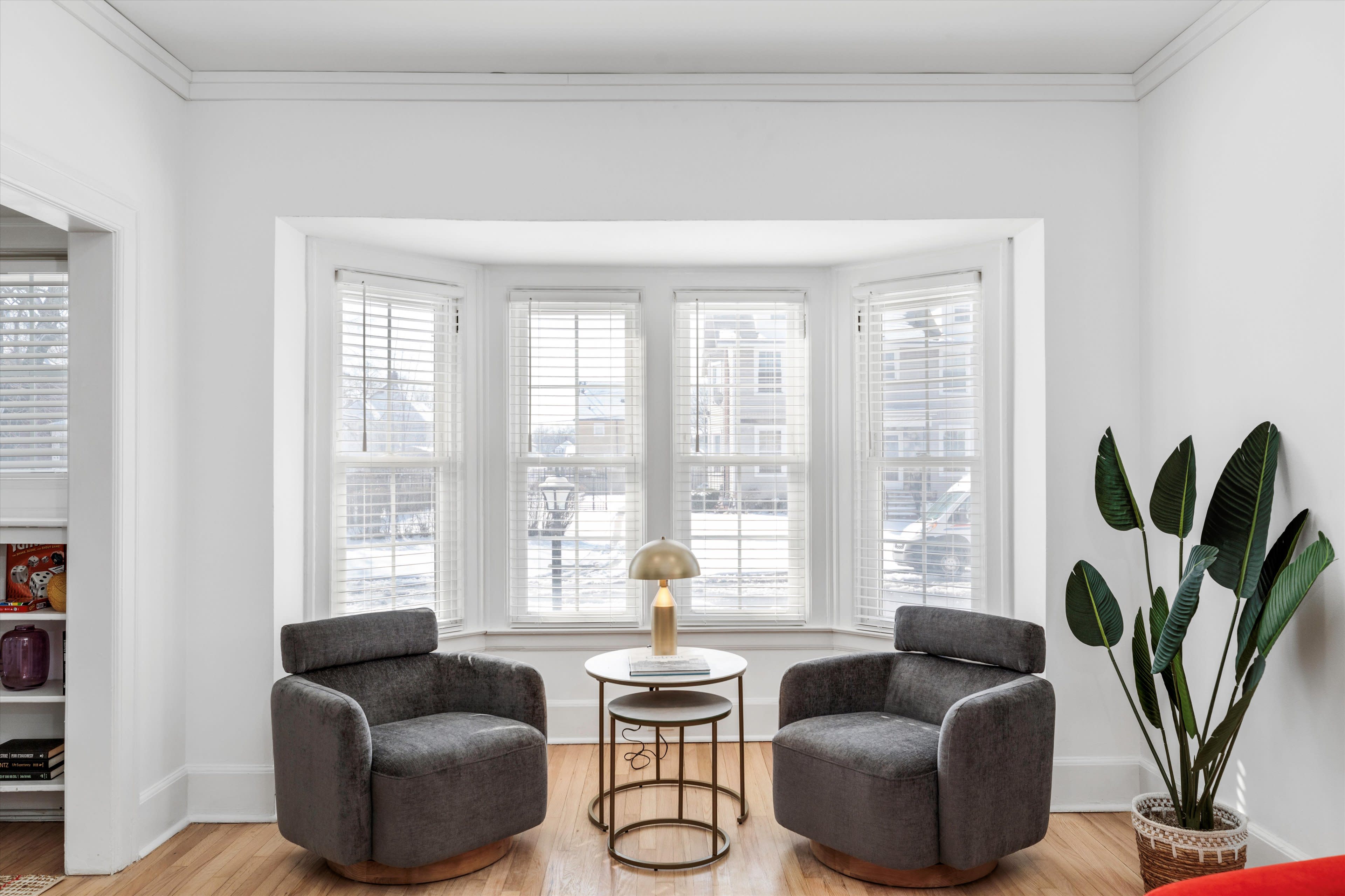 A cozy sitting area features two gray armchairs, a round coffee table, and a potted plant near a large bay window with blinds.