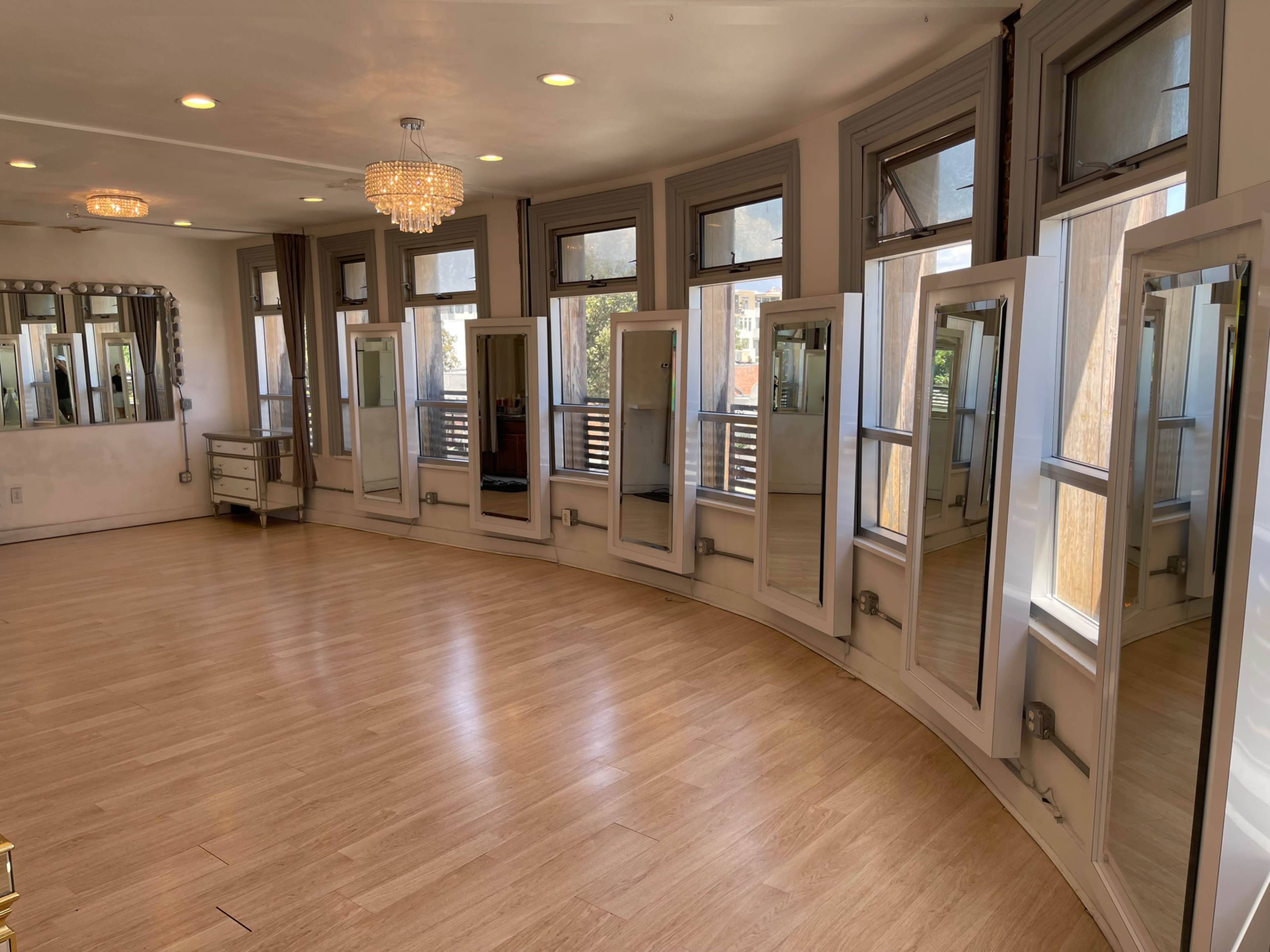 The image shows a spacious, curved interior room with multiple tall mirrors and large windows letting in natural light.