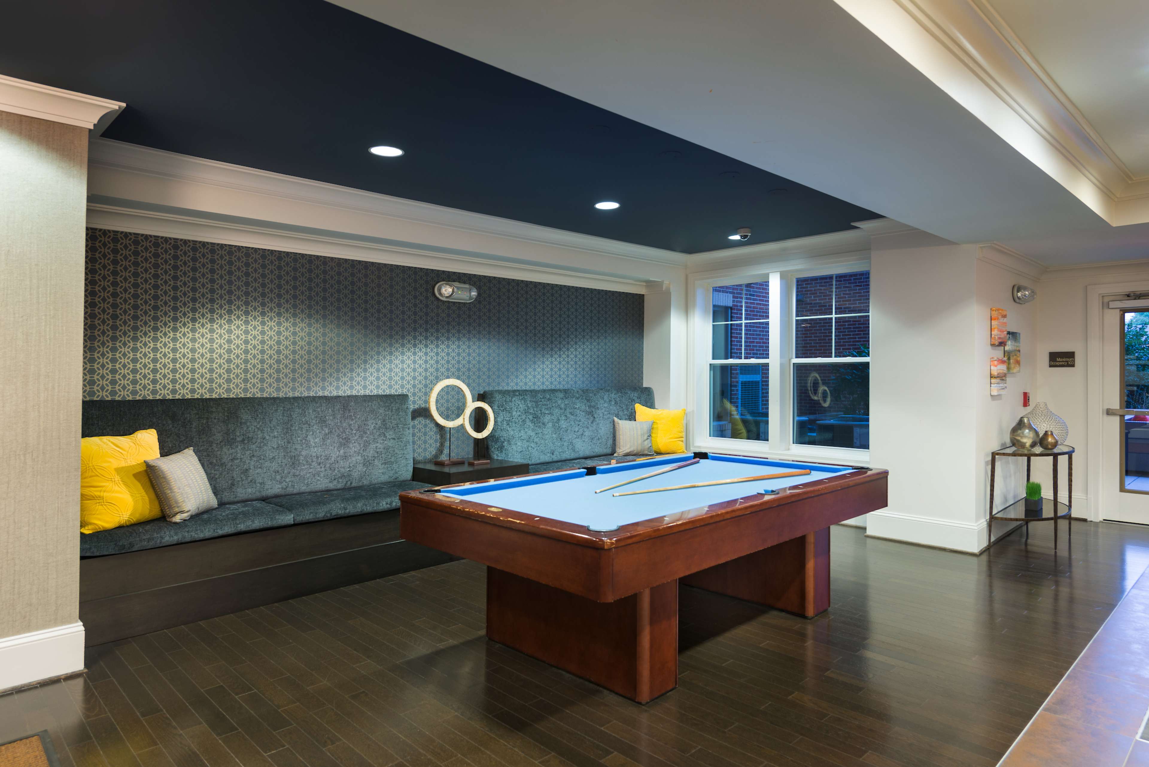 A billiards room featuring a blue pool table centered on a dark wooden floor, with a cushioned seating area along one wall and decorative elements on display.
