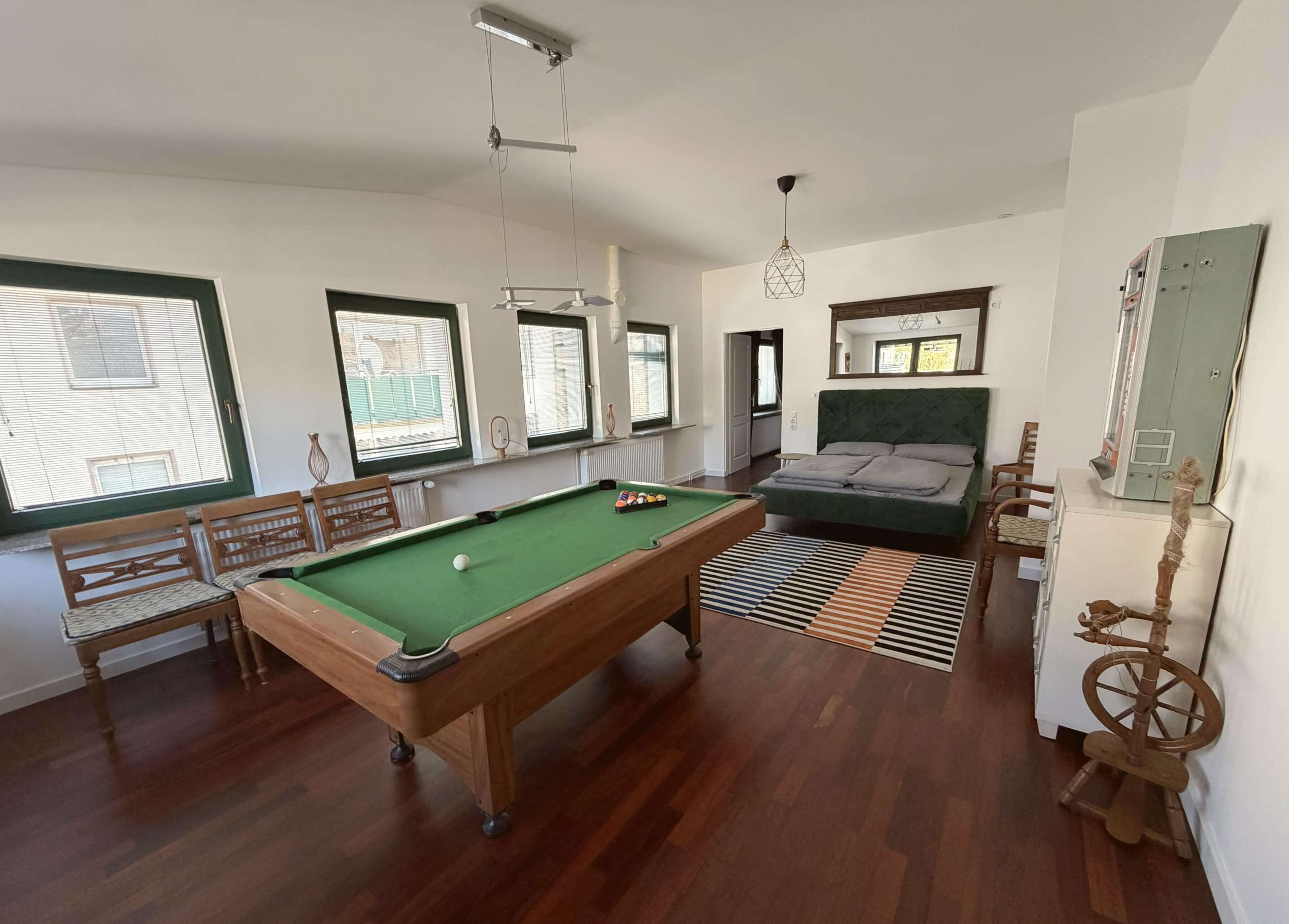 The image shows a room with a green billiards table in the center, surrounded by wooden chairs and a seating area with a bed and a mirror.