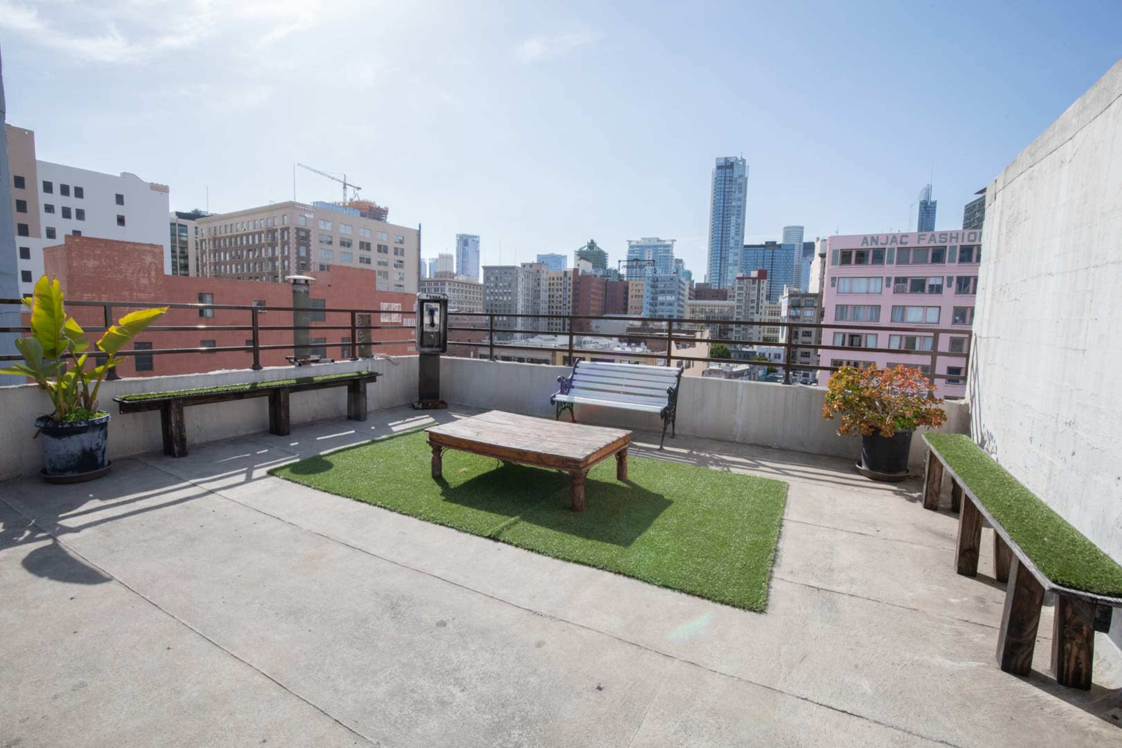 A rooftop terrace features wooden benches and a table on grassy flooring with a city skyline backdrop.