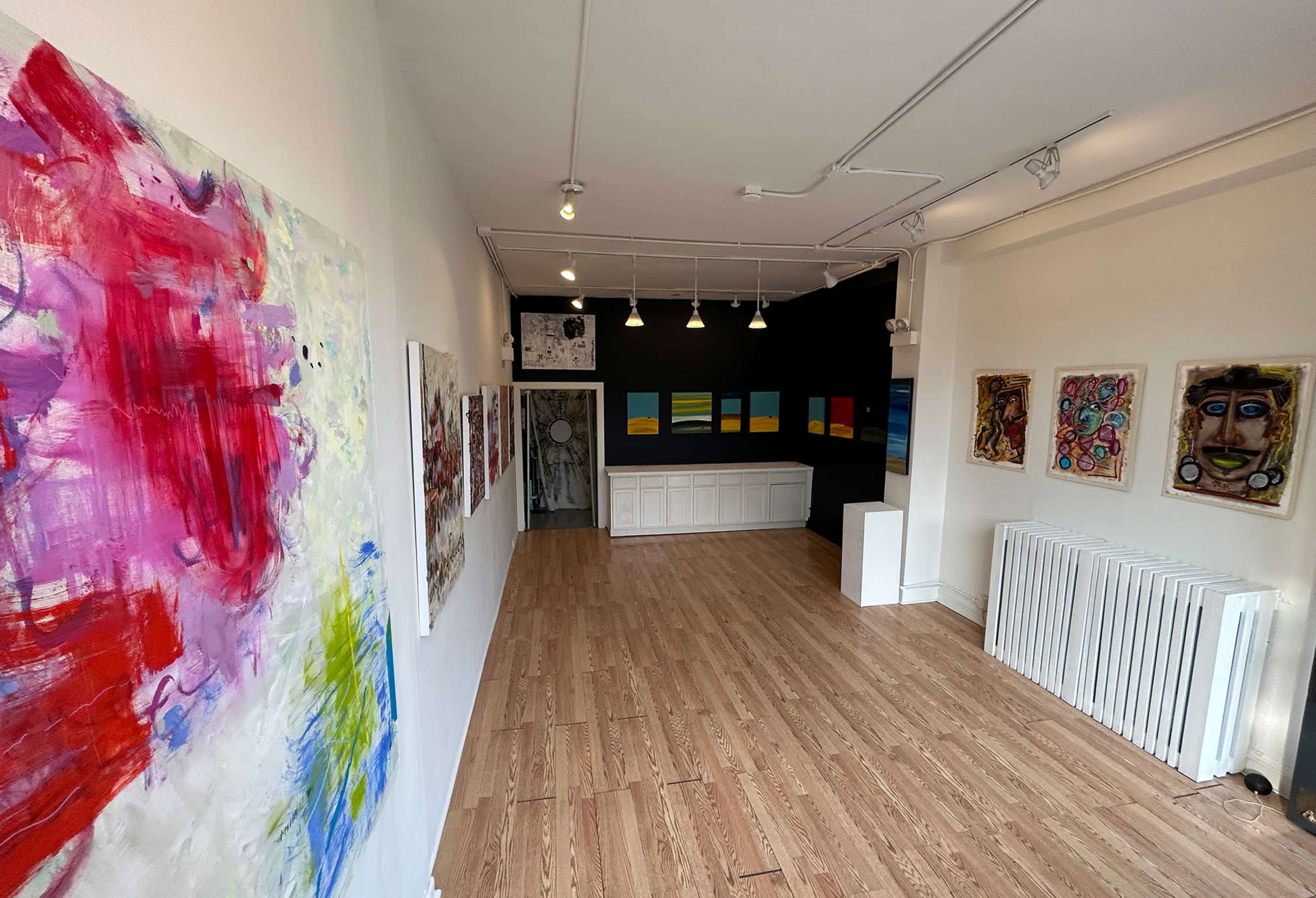 The image shows an art gallery interior featuring colorful paintings on the walls and a minimalist layout with wooden flooring and overhead lighting.