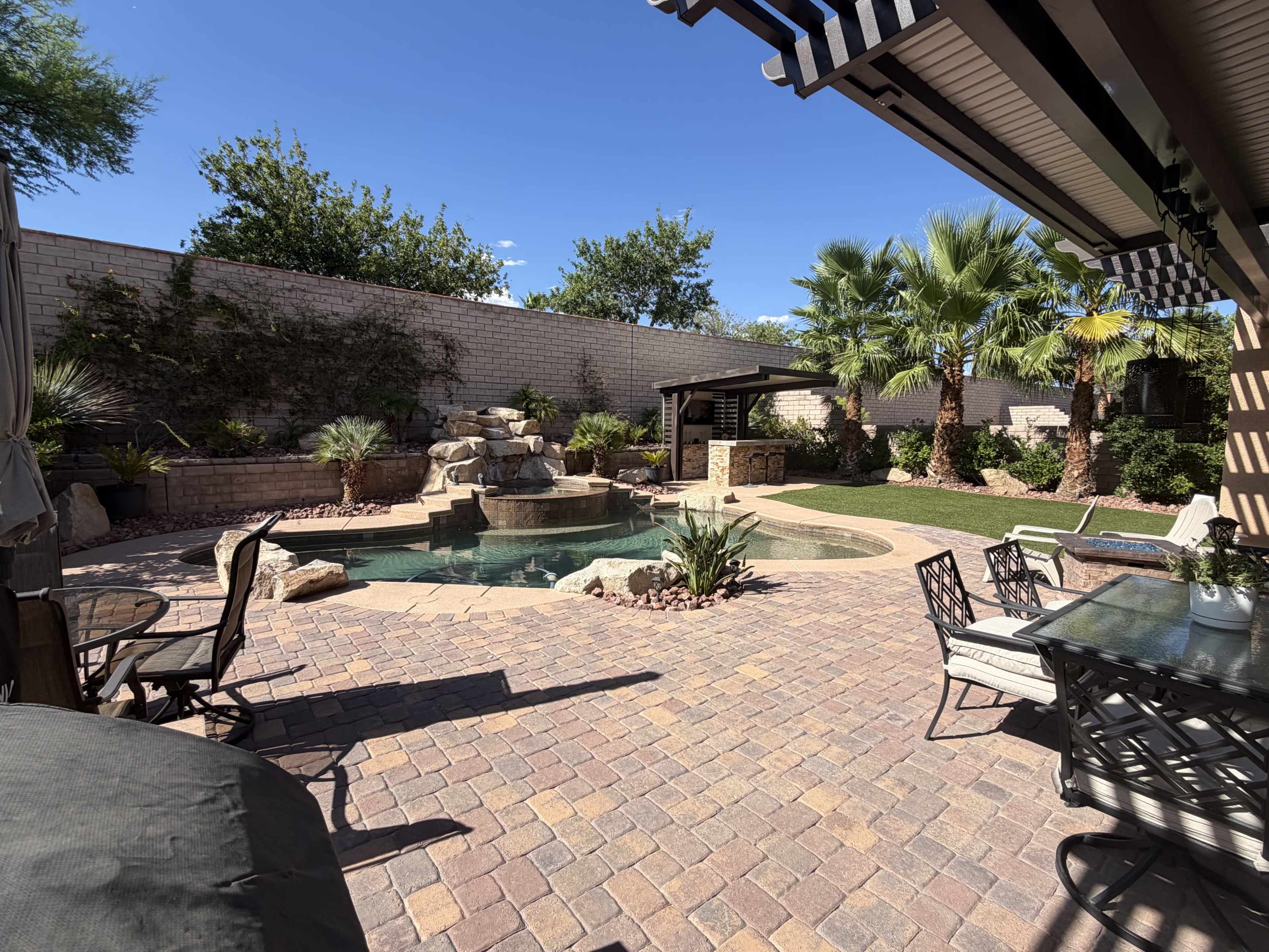 The image shows a landscaped backyard with a swimming pool, rock features, palm trees, and a patio area with outdoor furniture.