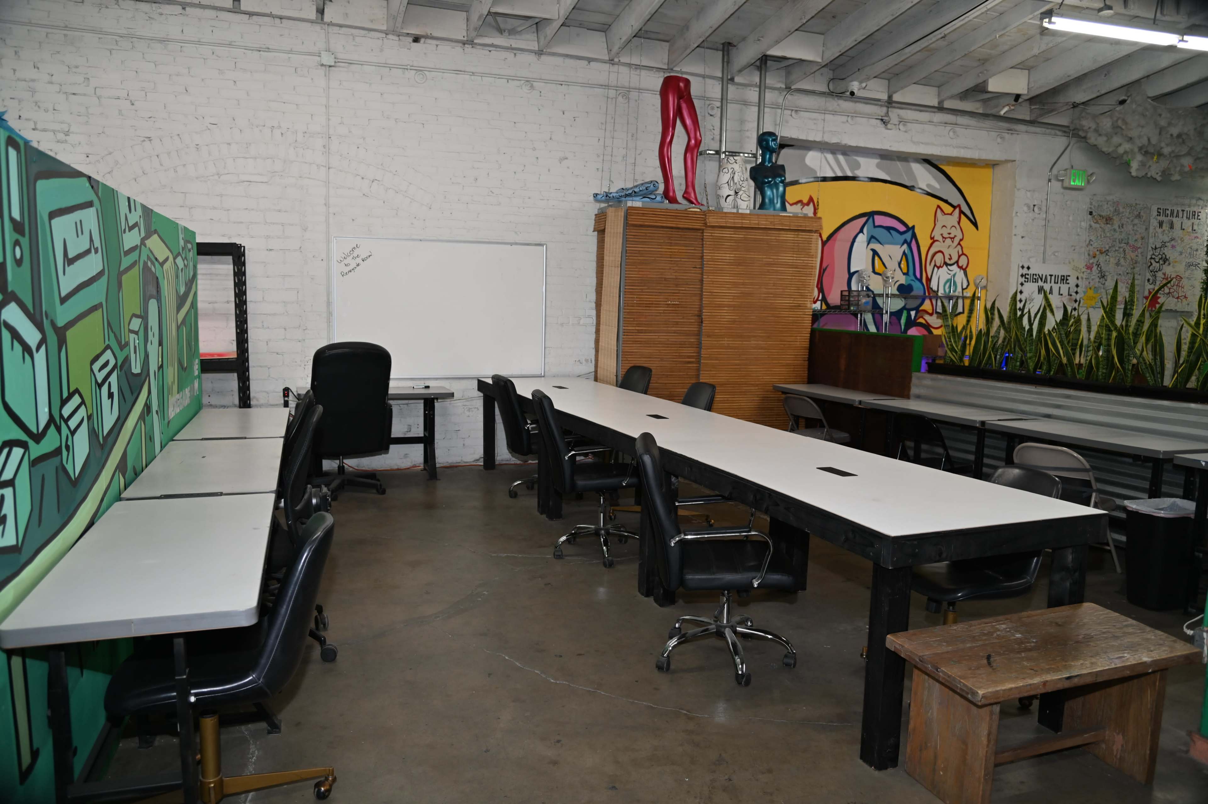 The image shows a spacious, industrial-style office with several black office chairs, a long white table, and colorful wall murals.