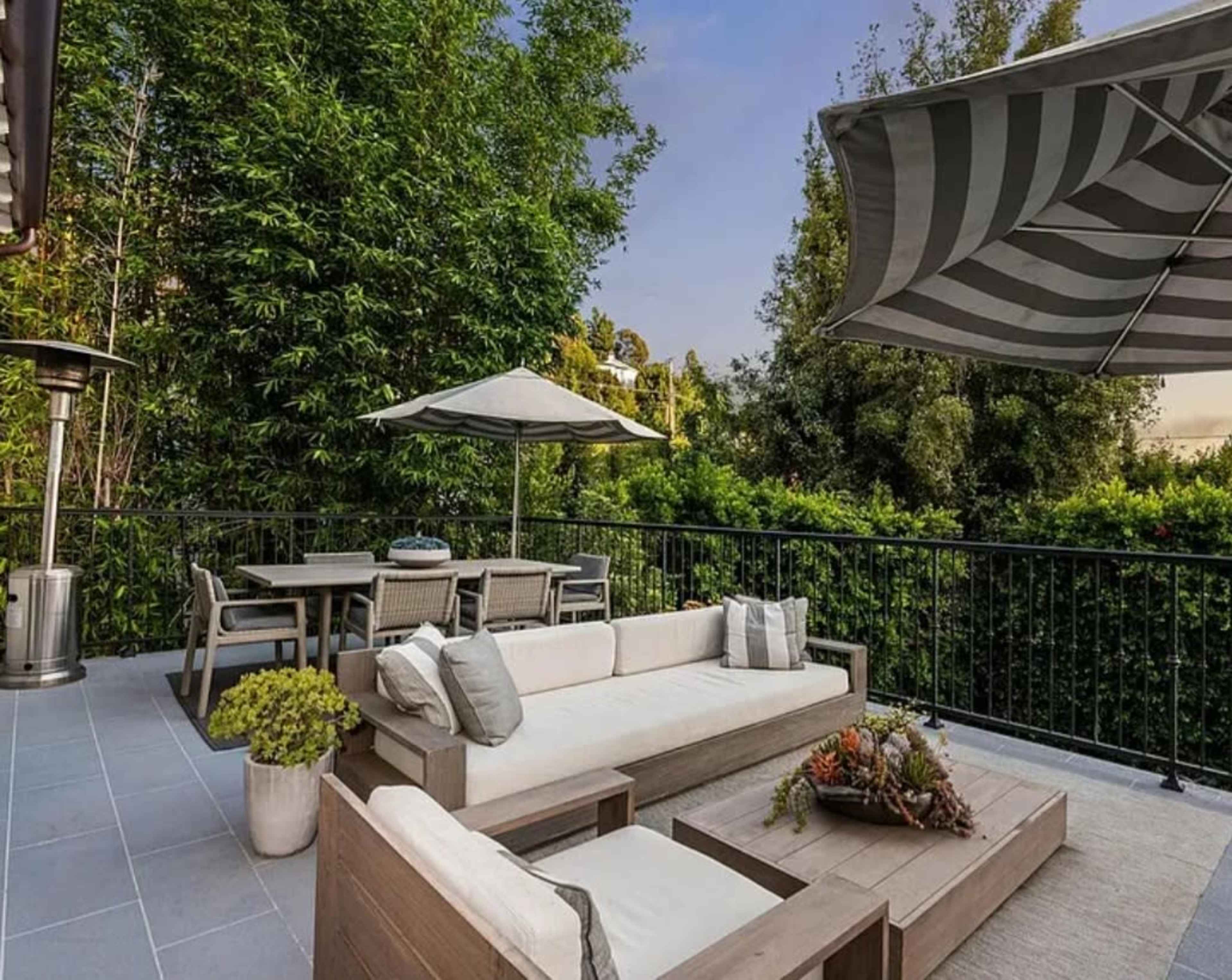 A modern outdoor patio features a wooden seating arrangement, a dining table with chairs, and an umbrella, surrounded by lush greenery.