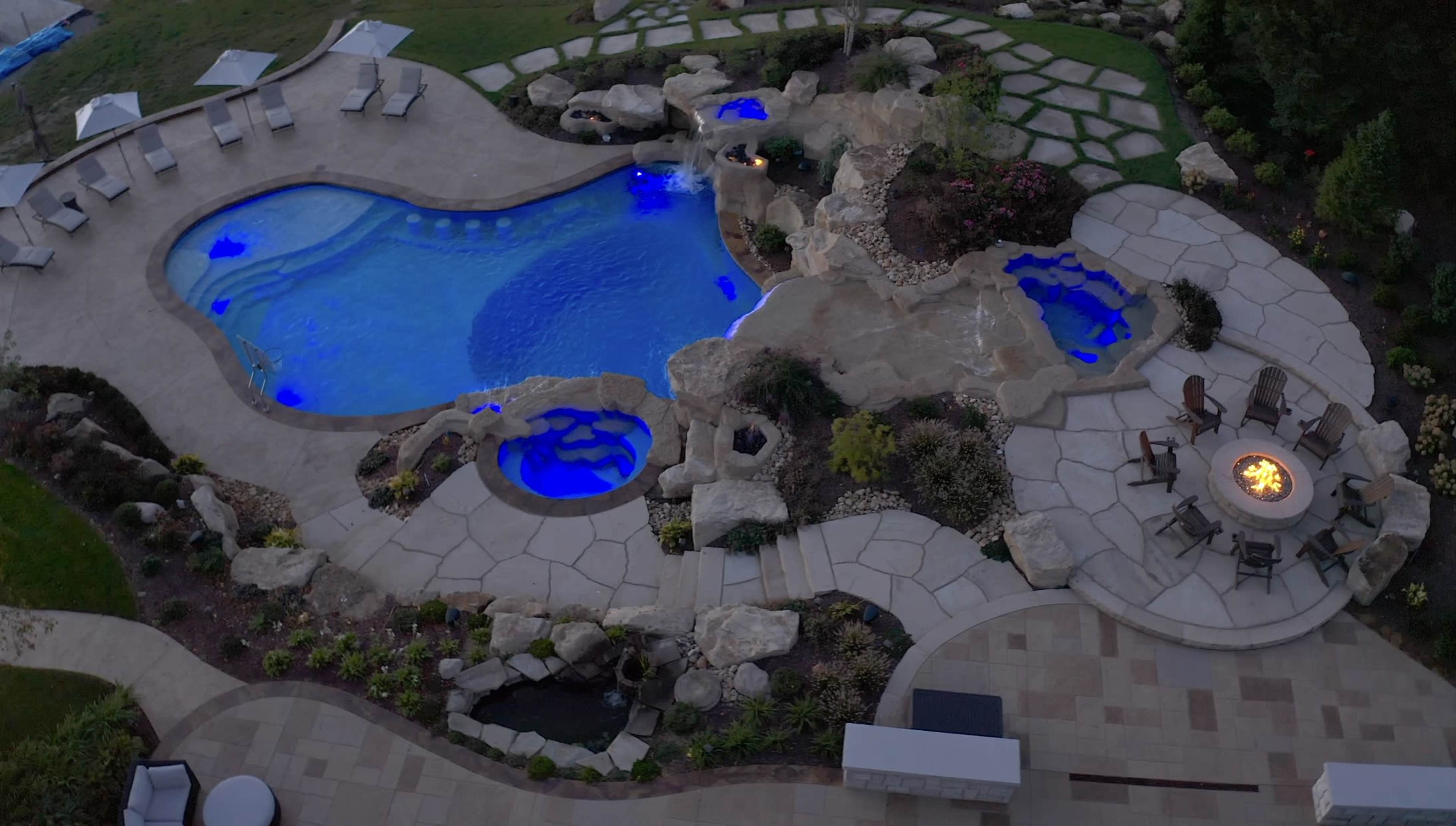 The image shows an aerial view of a landscaped backyard with a swimming pool, spa areas, and a fire pit surrounded by rocks and vegetation.
