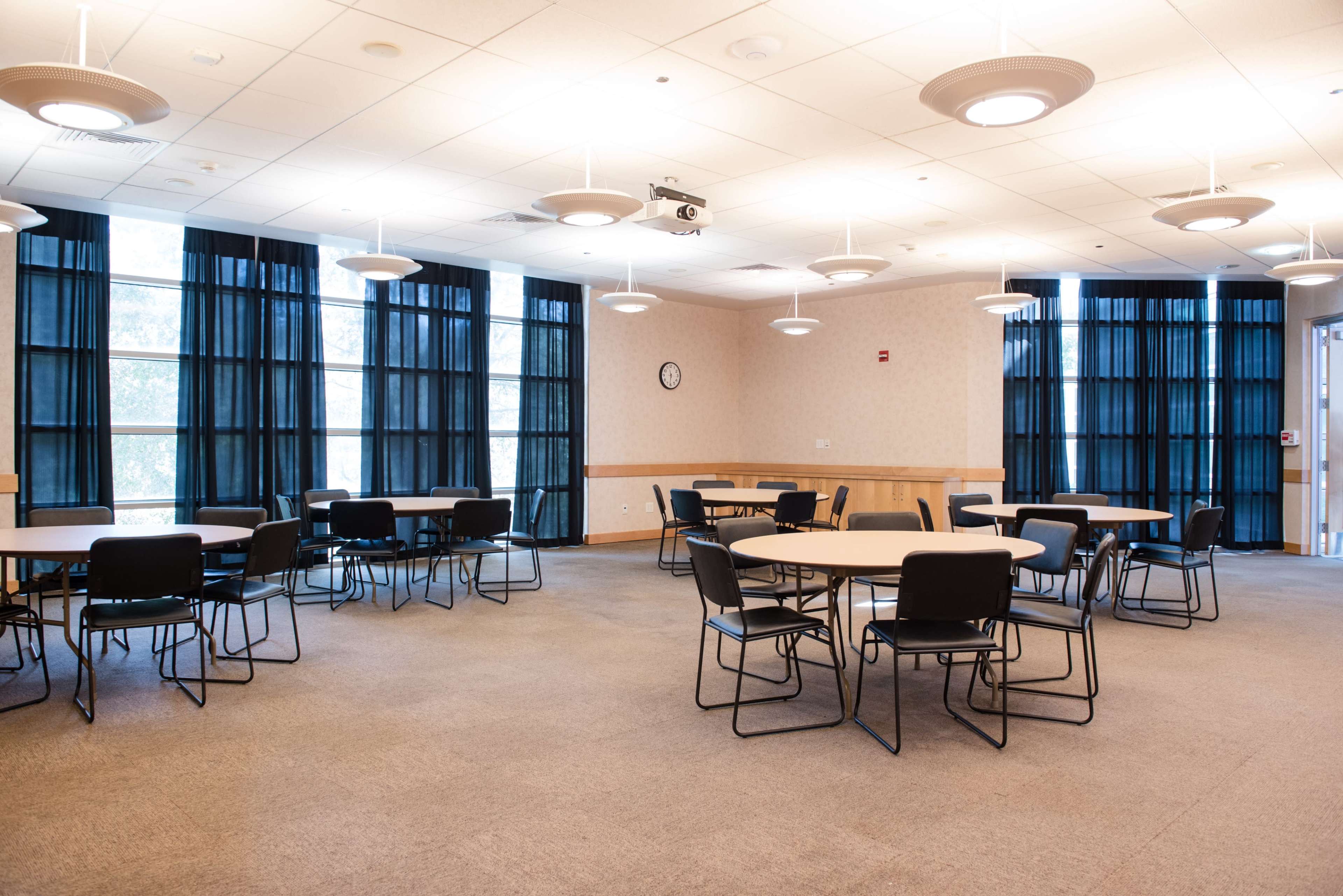A conference room featuring several round tables and chairs with large windows covered by dark curtains.