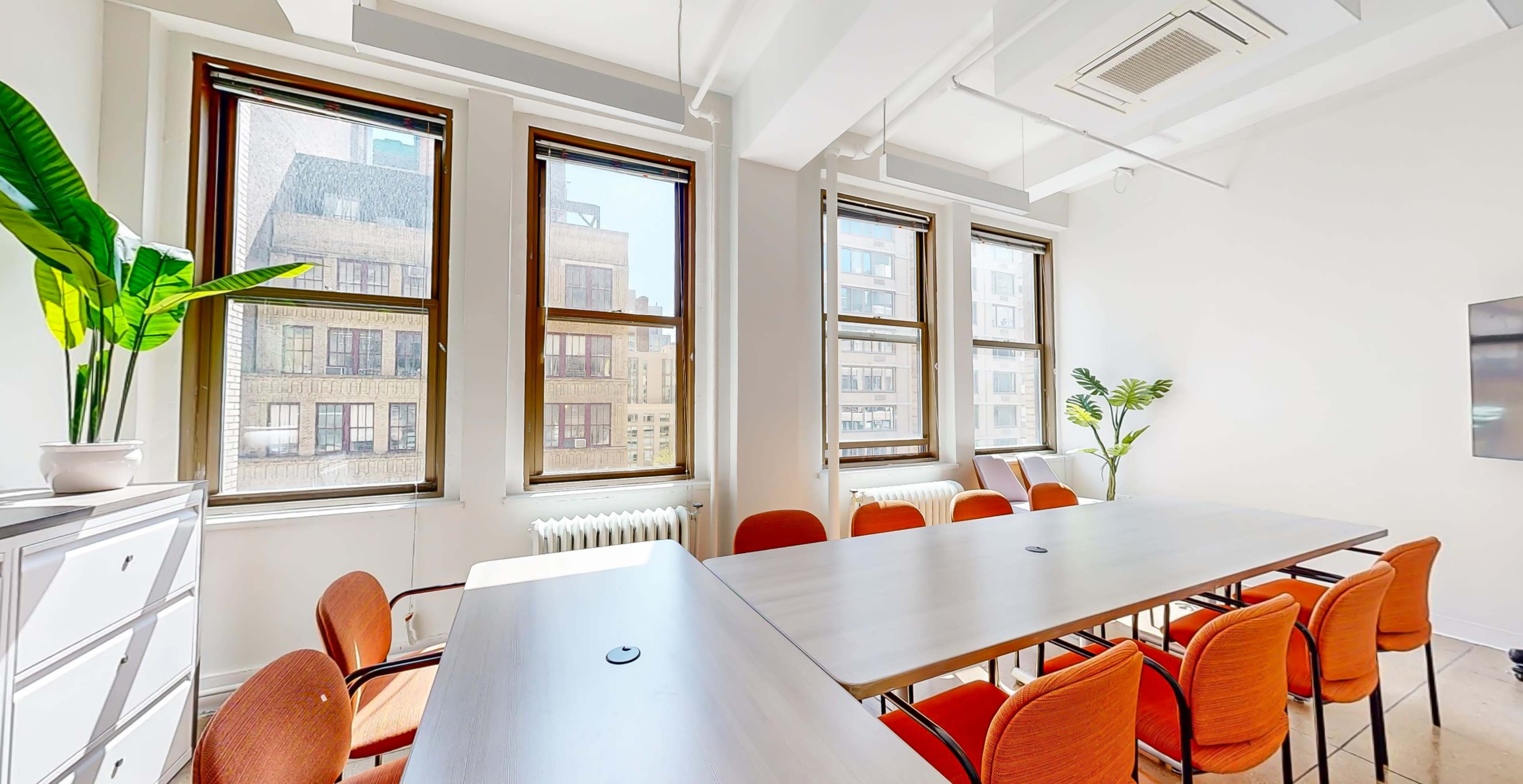 A bright meeting room features large windows, a long table, and orange chairs arranged around it.
