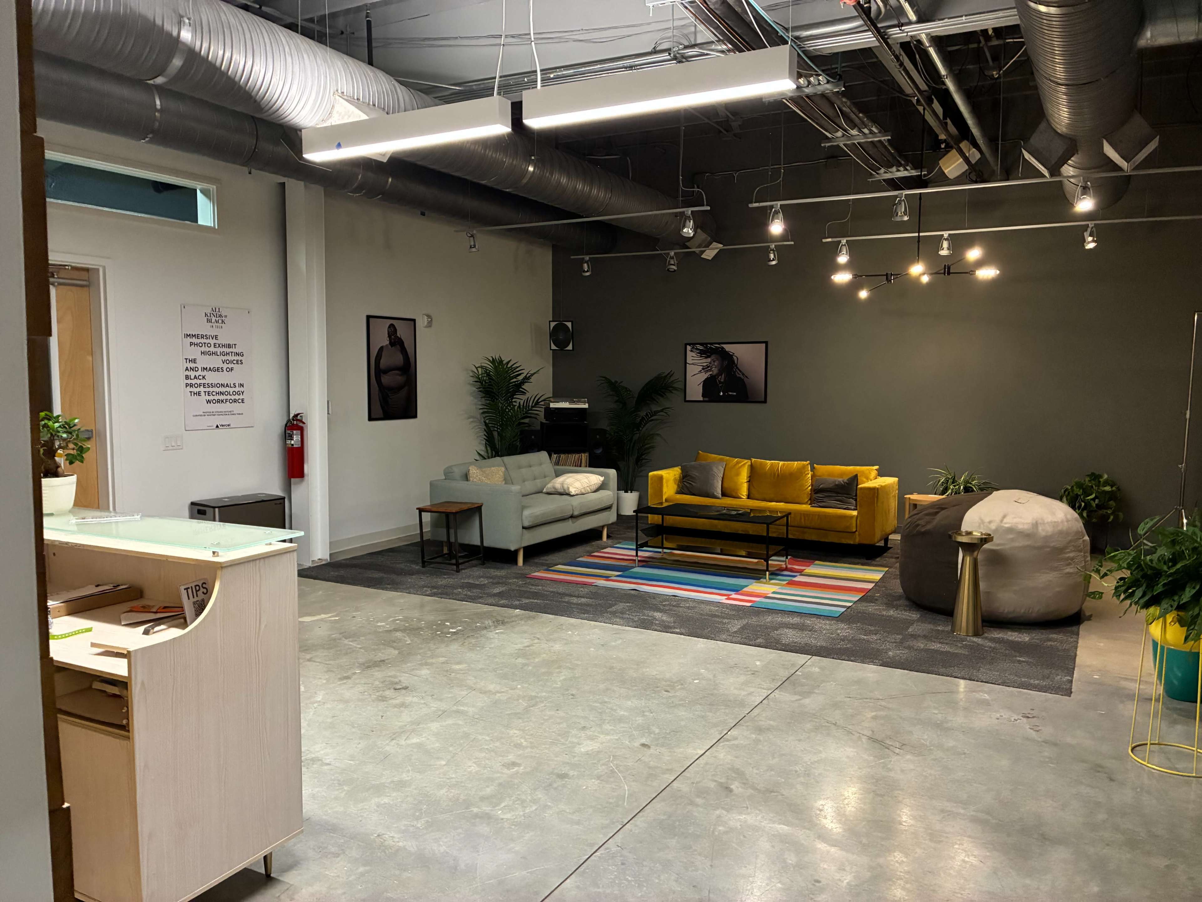 The image shows a modern office lounge area with a mix of seating options, including a gray sofa, a yellow couch, and a bean bag, placed on a colorful rug against a gray wall.