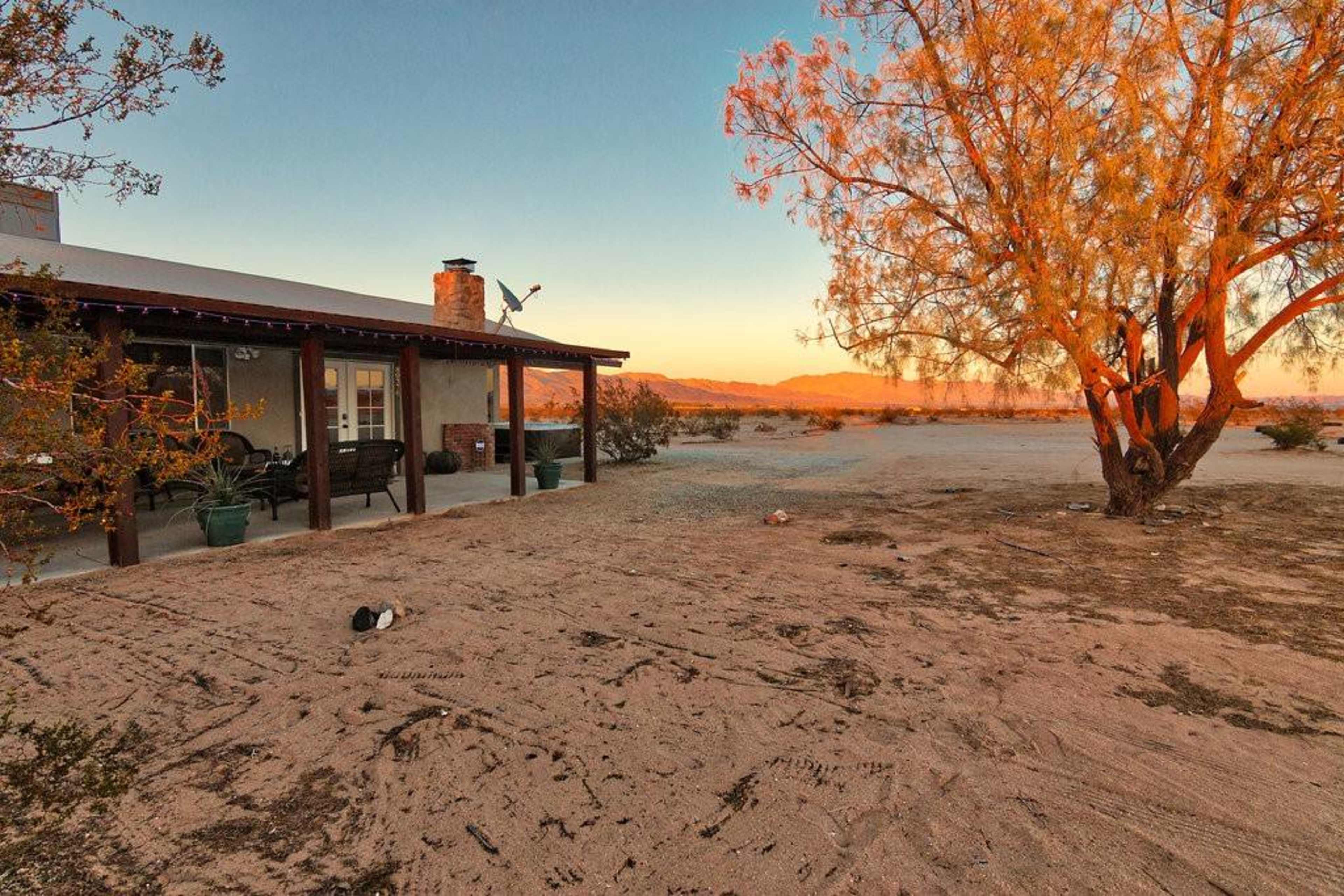 A single-story house with a porch is surrounded by desert landscape and a large tree at sunset.