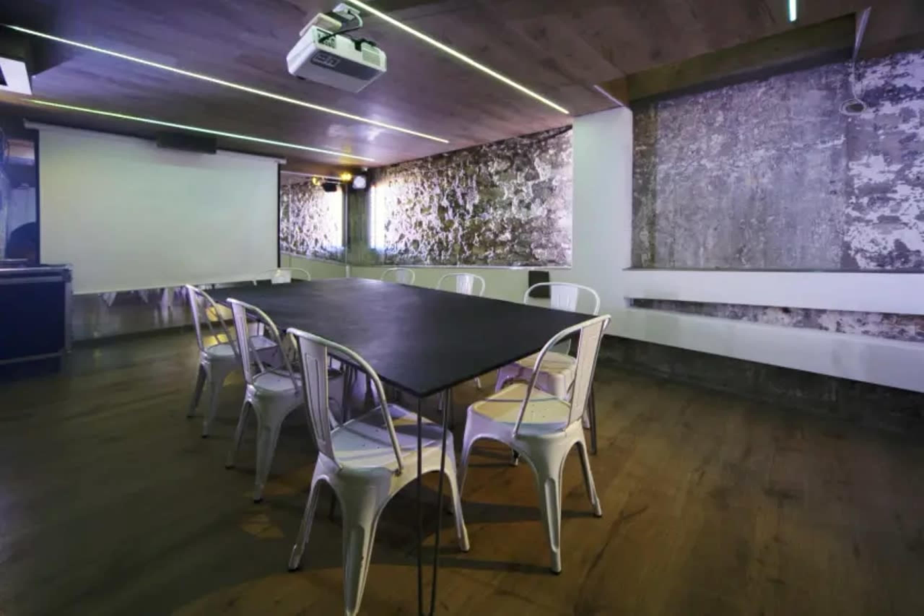 A rectangular black table surrounded by metal chairs is positioned in a modern meeting room with exposed concrete walls and a projector on the ceiling.