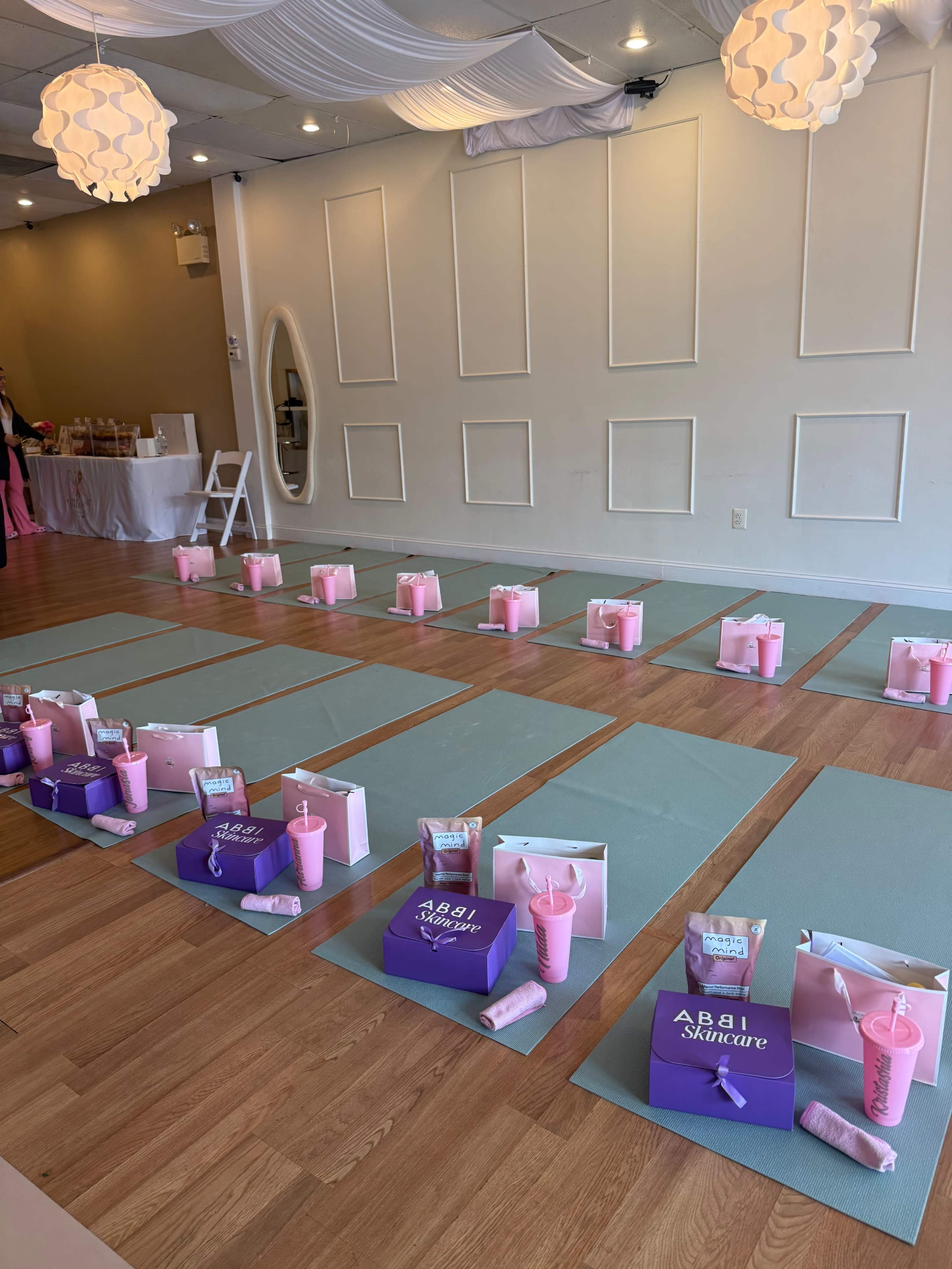 The image shows a yoga studio set up with mats arranged in rows, each accompanied by pink-themed gift bags and cups.