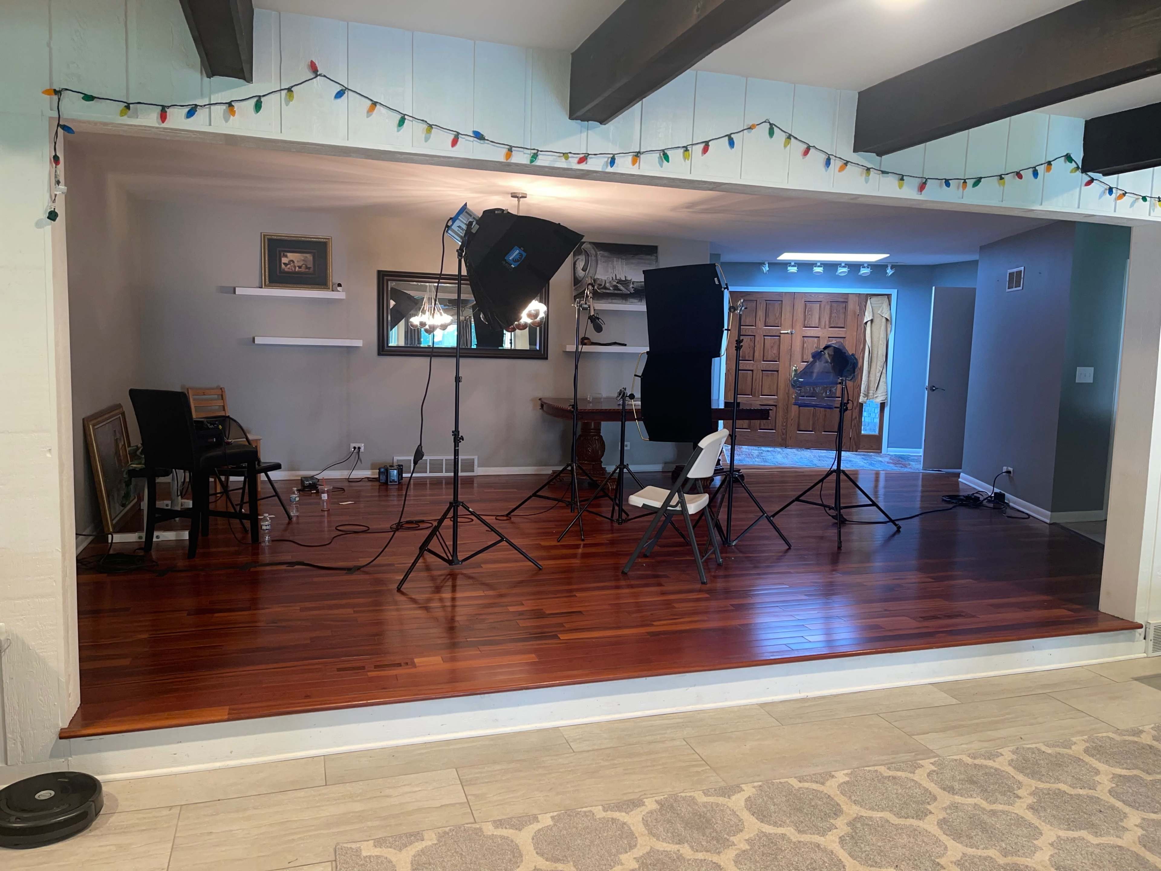 A studio setup with lighting equipment and chairs in a section of a room featuring hardwood floors and a doorway leading to another area.