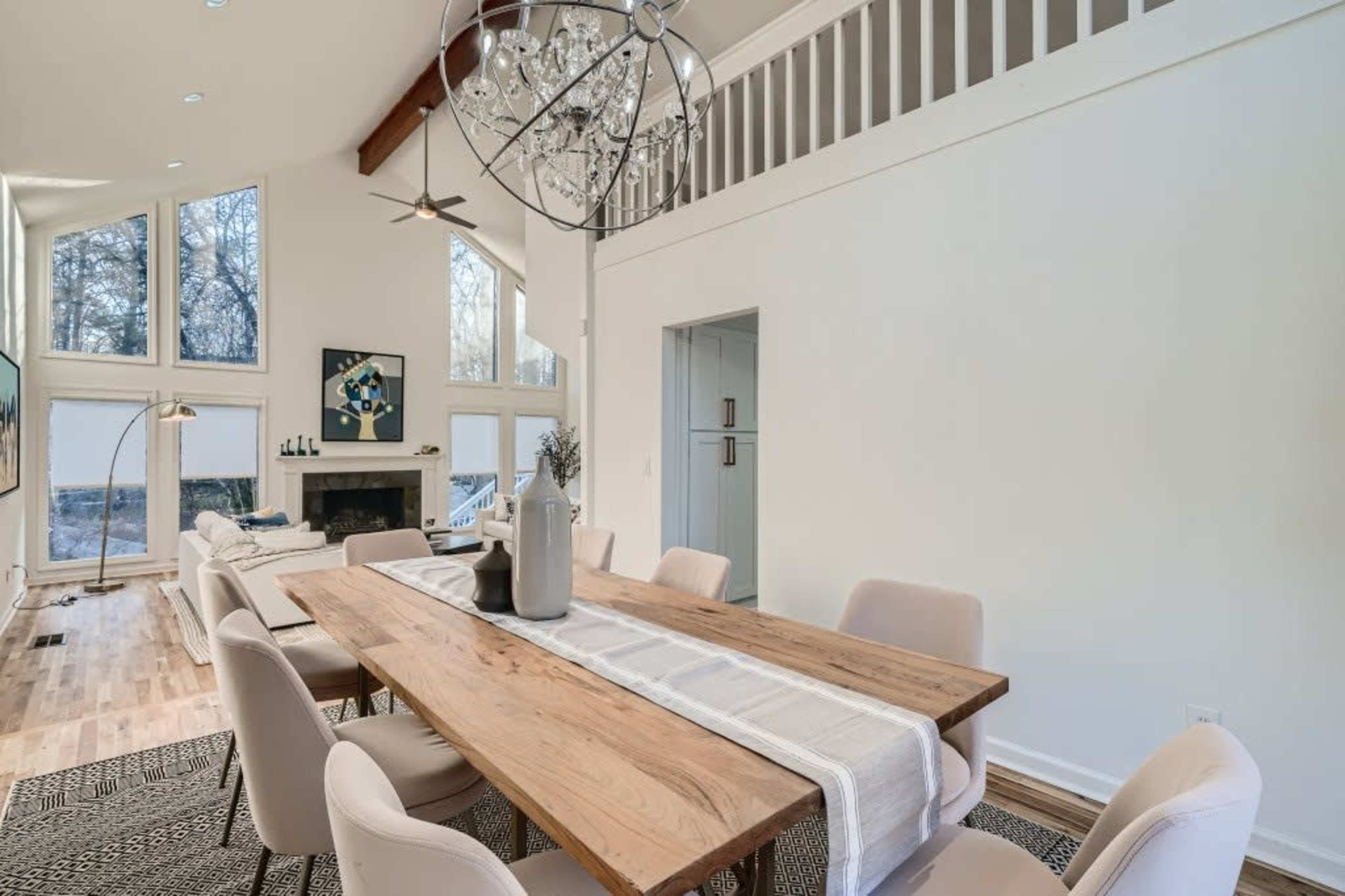 A spacious dining area featuring a long wooden table surrounded by upholstered chairs, with a chandelier above and large windows letting in natural light.