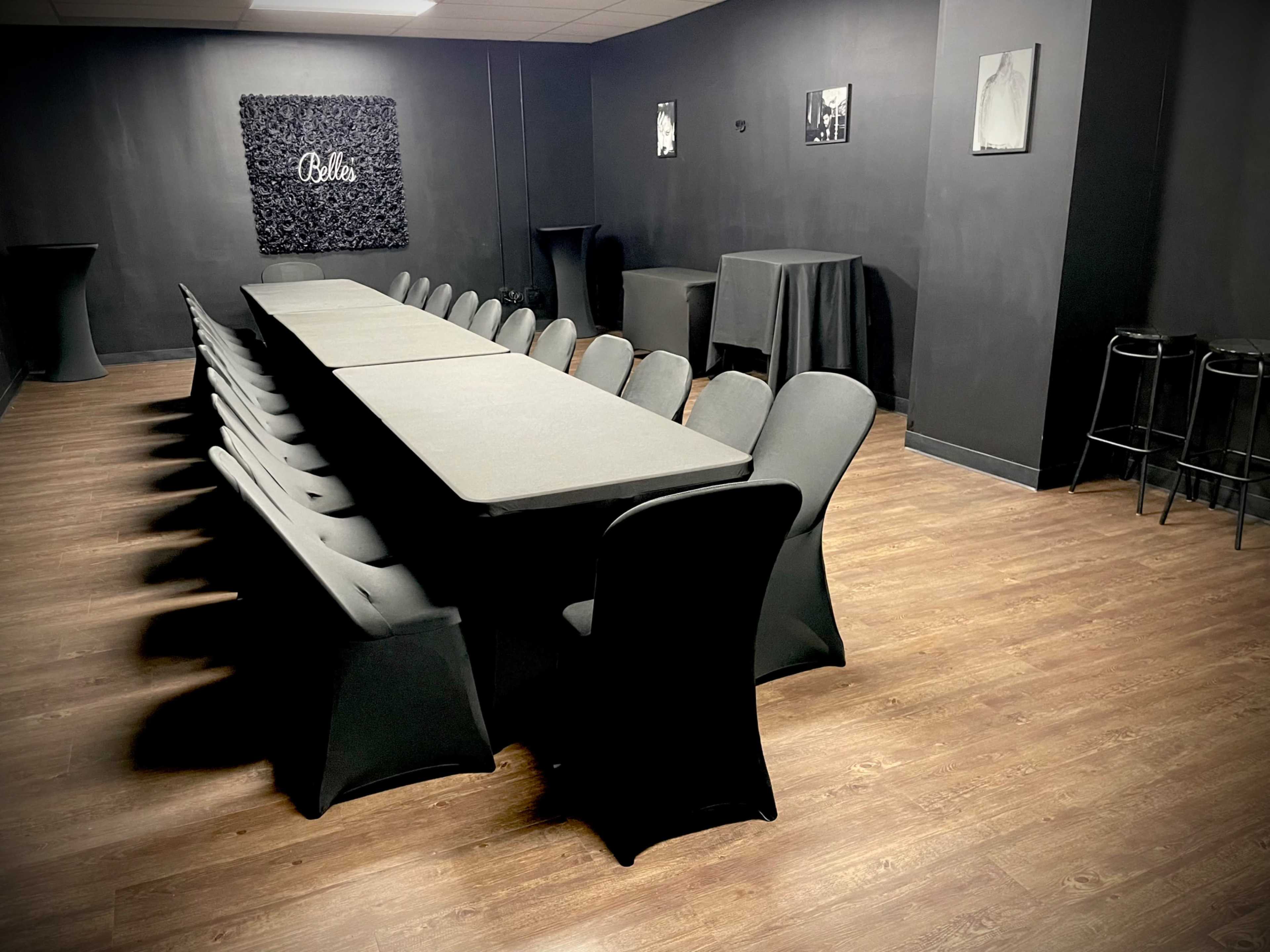 A long rectangular table with black tablecloths is arranged in a dimly lit room, surrounded by chairs, with black walls and a decorative wall featuring the word "Bella."