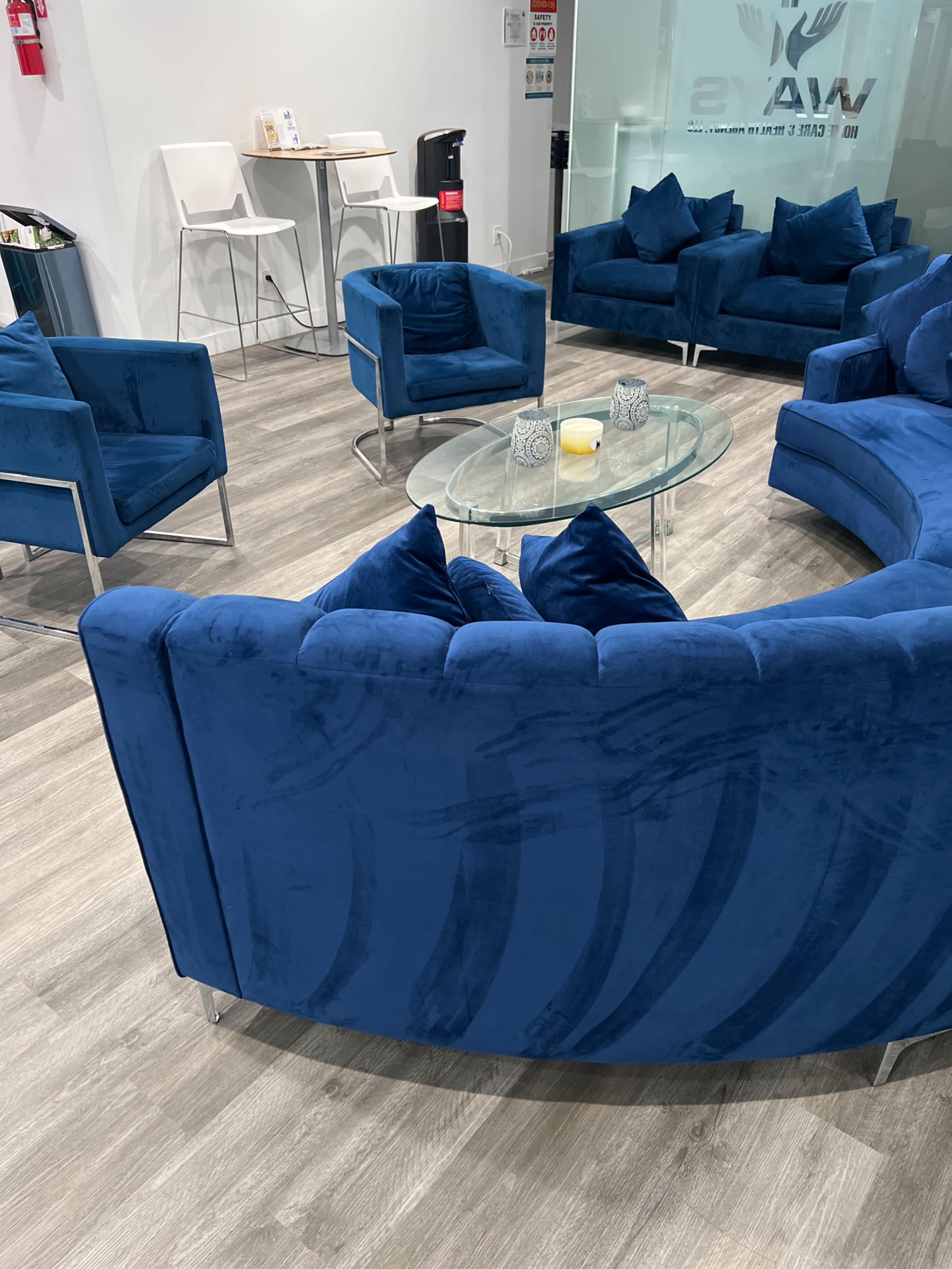 A modern seating area featuring a circular arrangement of blue upholstered sofas and chairs around a glass coffee table in a well-lit room.