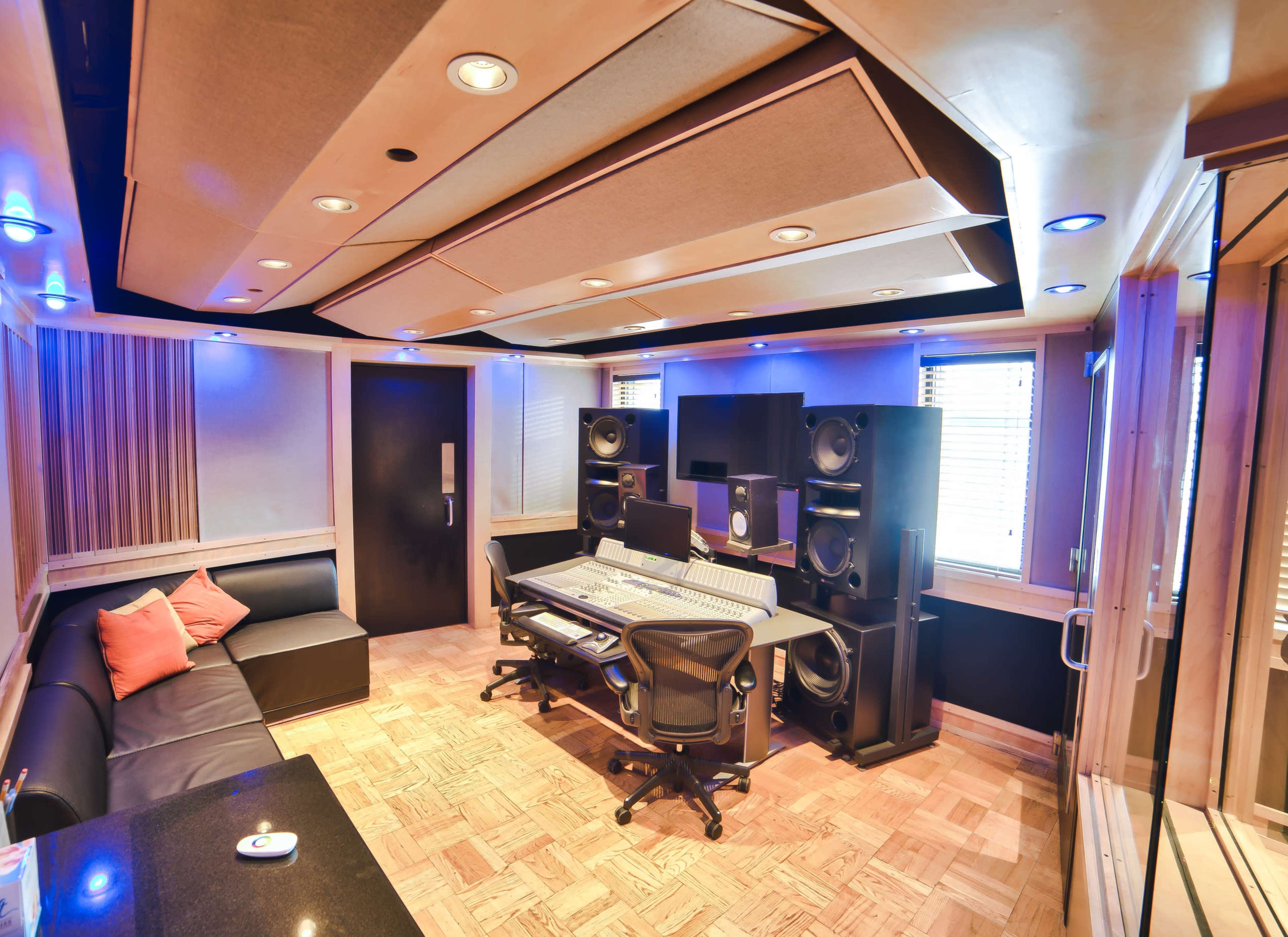 The image shows a modern music production studio featuring a central mixing console, large speakers, and comfortable seating.