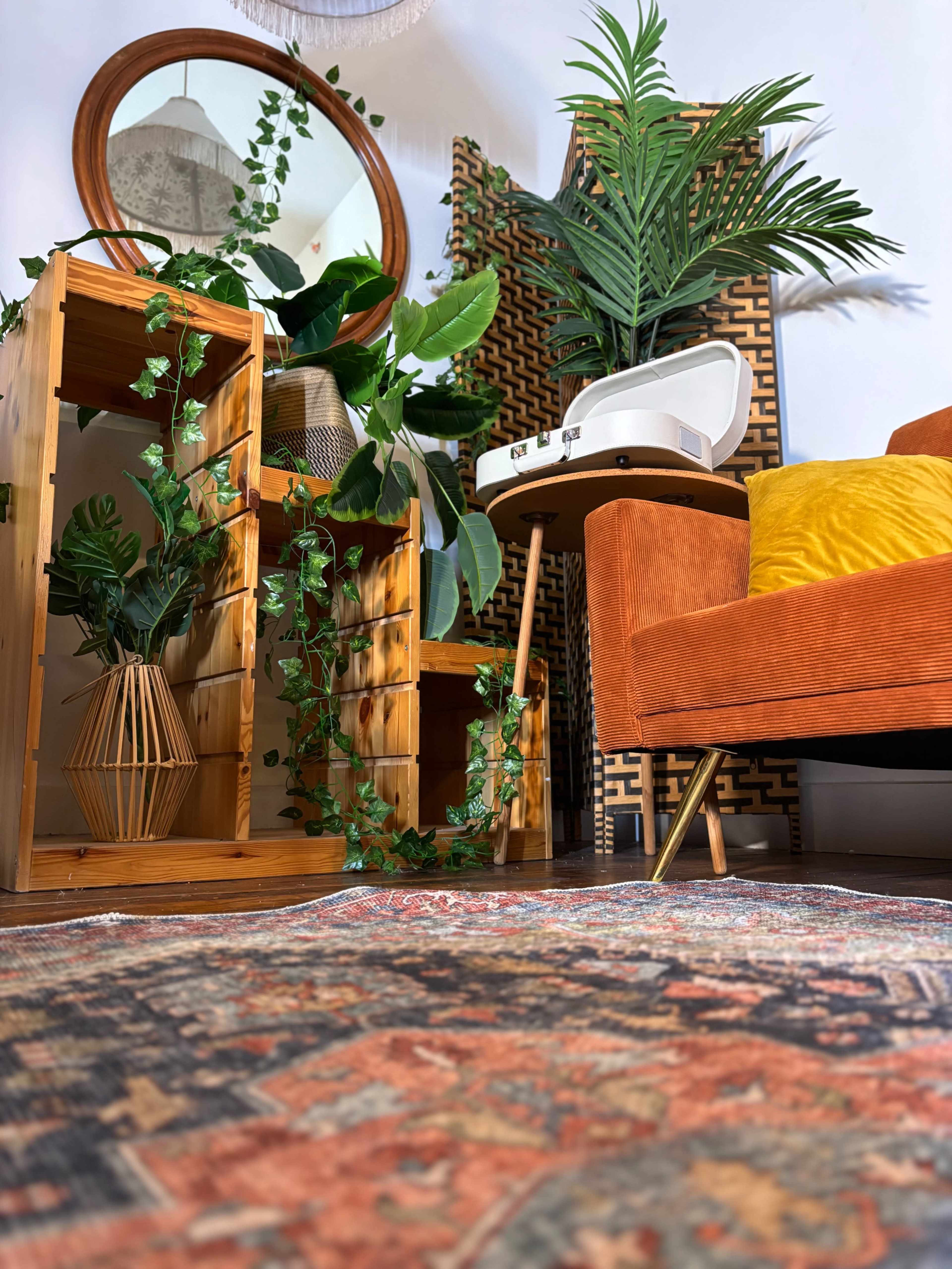 The image shows a cozy interior space featuring an orange chair, a wooden shelf with plants, a round mirror, and a small table with a record player.