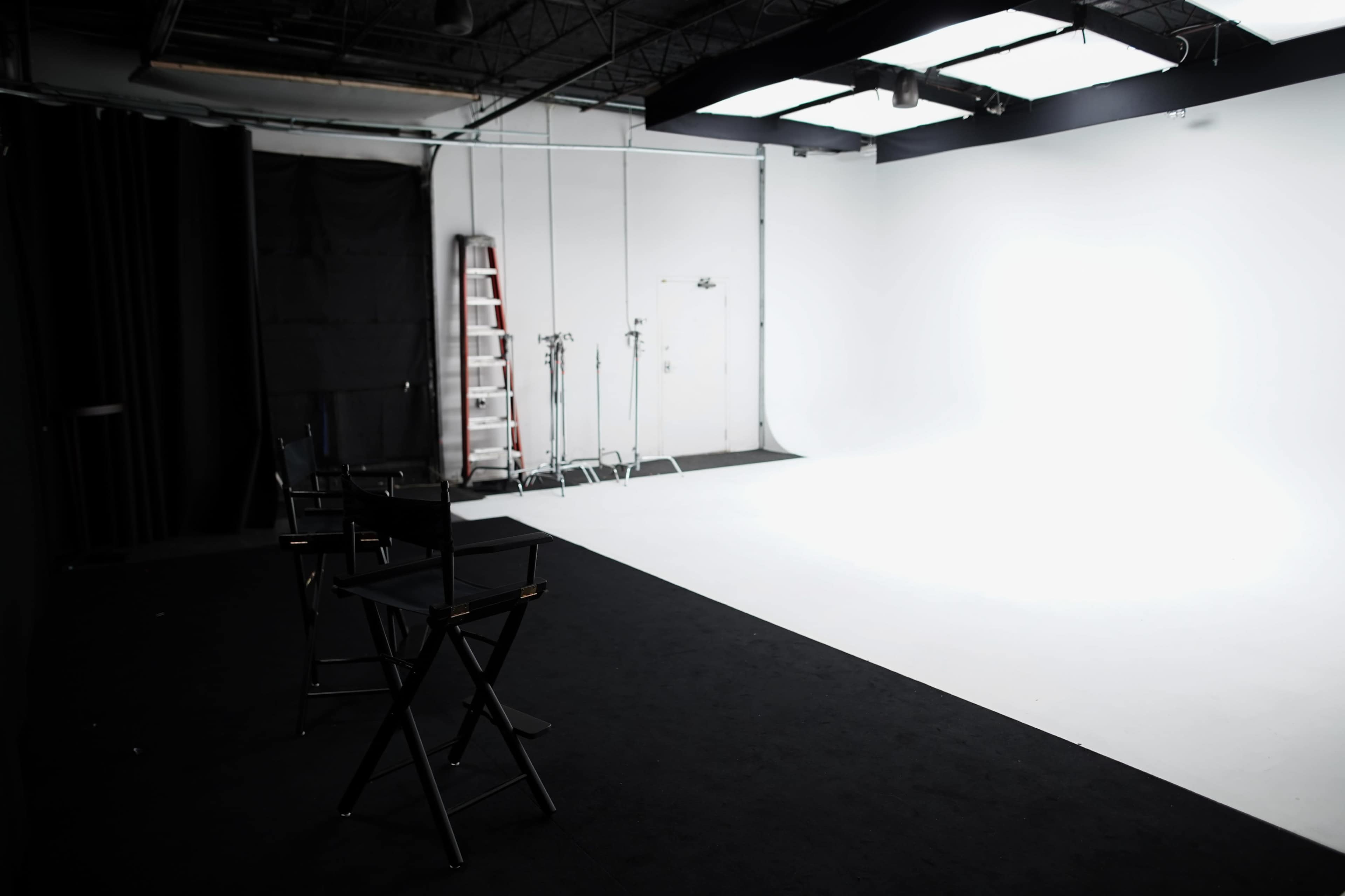 A photography studio features empty black director's chairs facing a blank white backdrop, with a ladder and lighting equipment in the background.