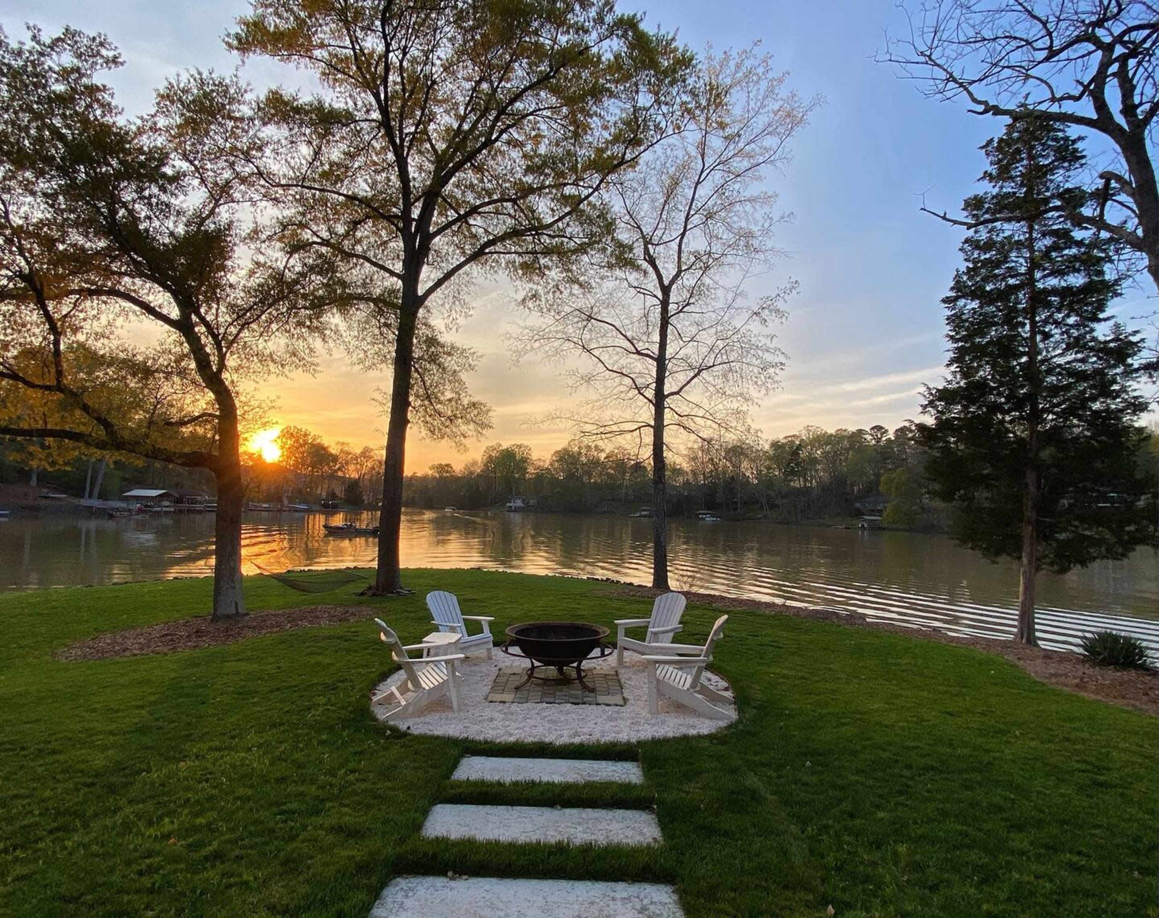 A fire pit surrounded by white chairs is situated on a grassy area by the water, with trees lining the shore and a sunset reflecting on the lake.