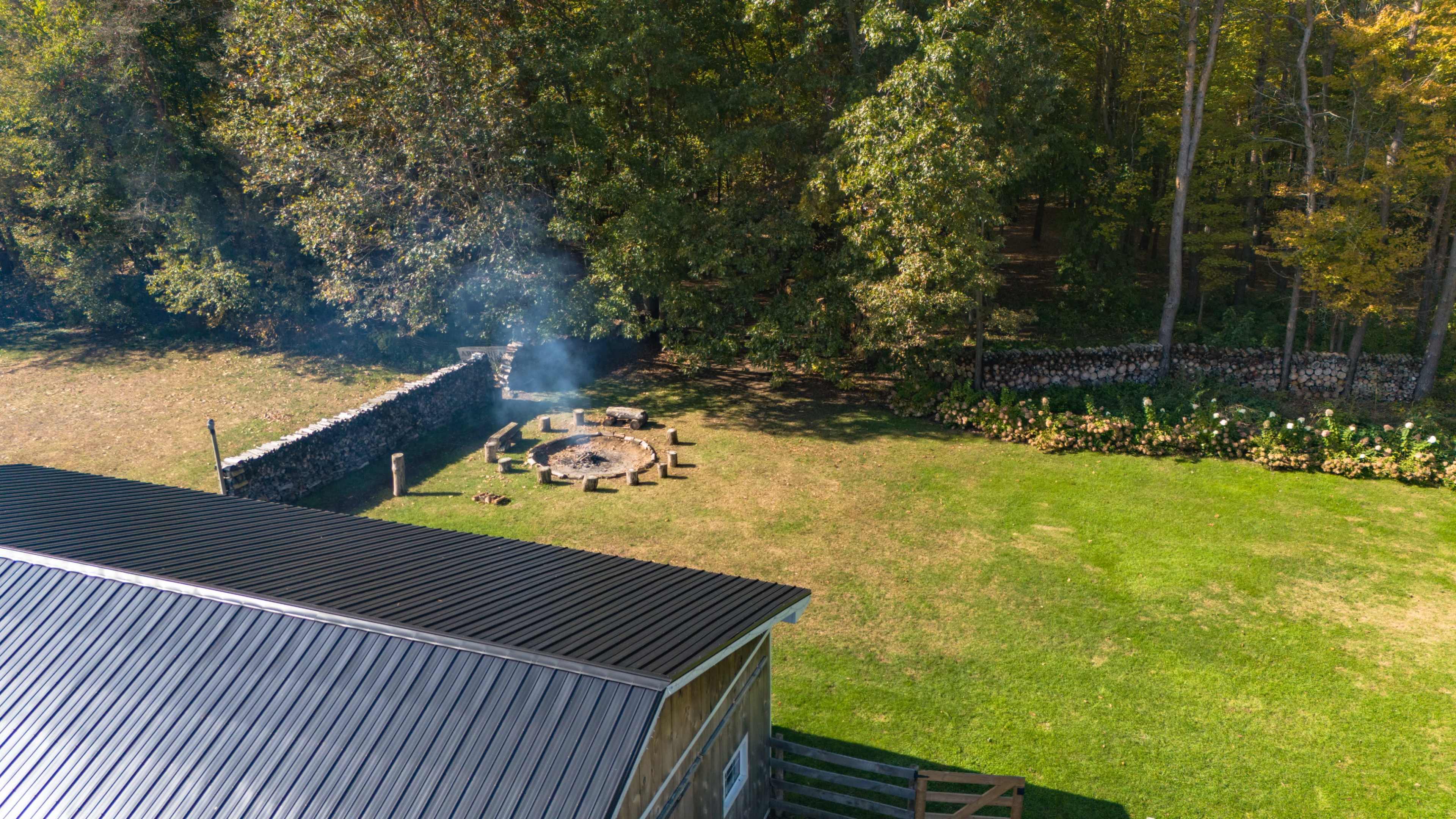 The image shows a circular fire pit surrounded by wooden seating, located on a grassy area next to a wooded backdrop.