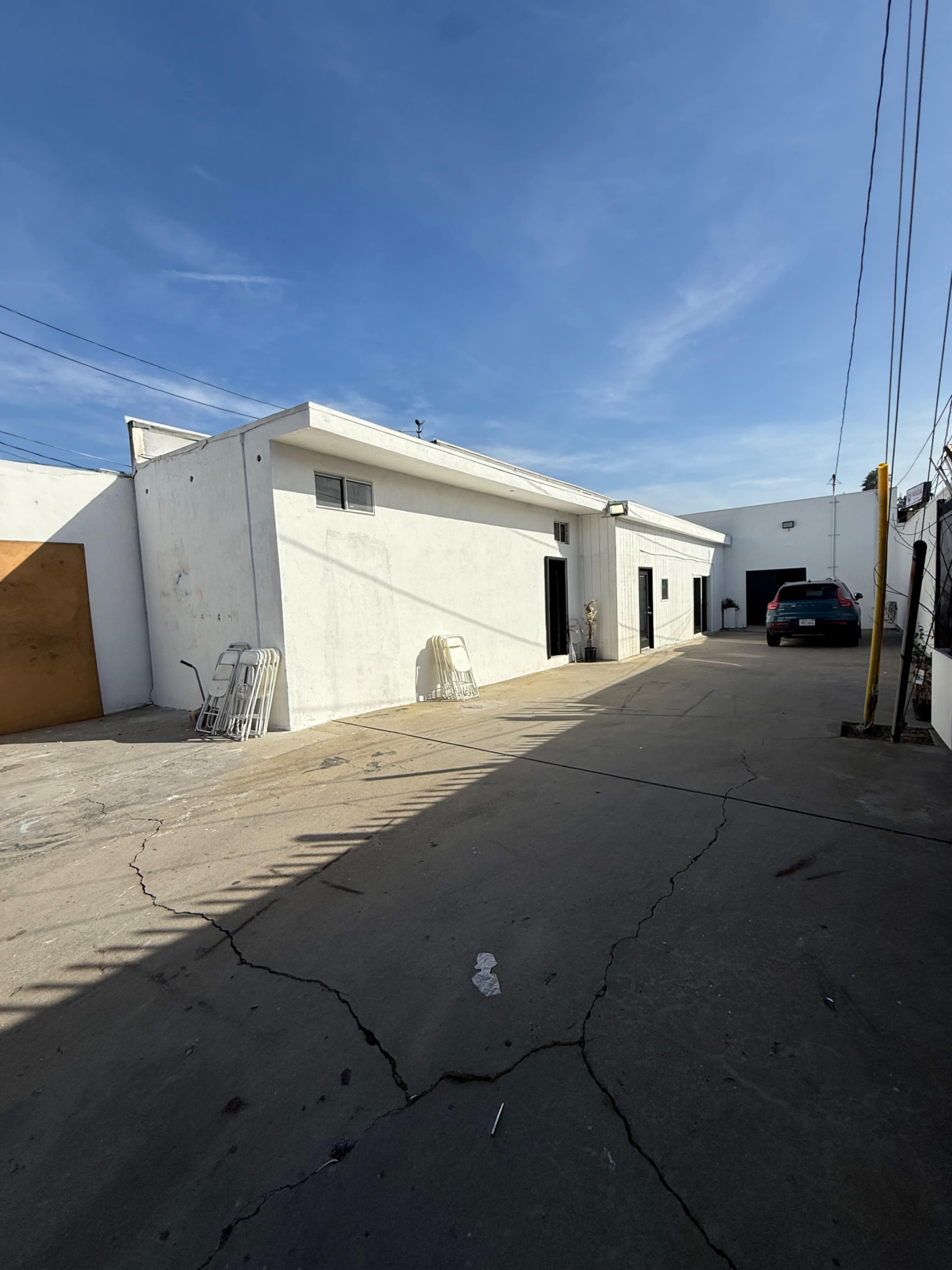 The image shows a narrow concrete alleyway beside a white building, with a parked car and metal folding chairs against the wall.