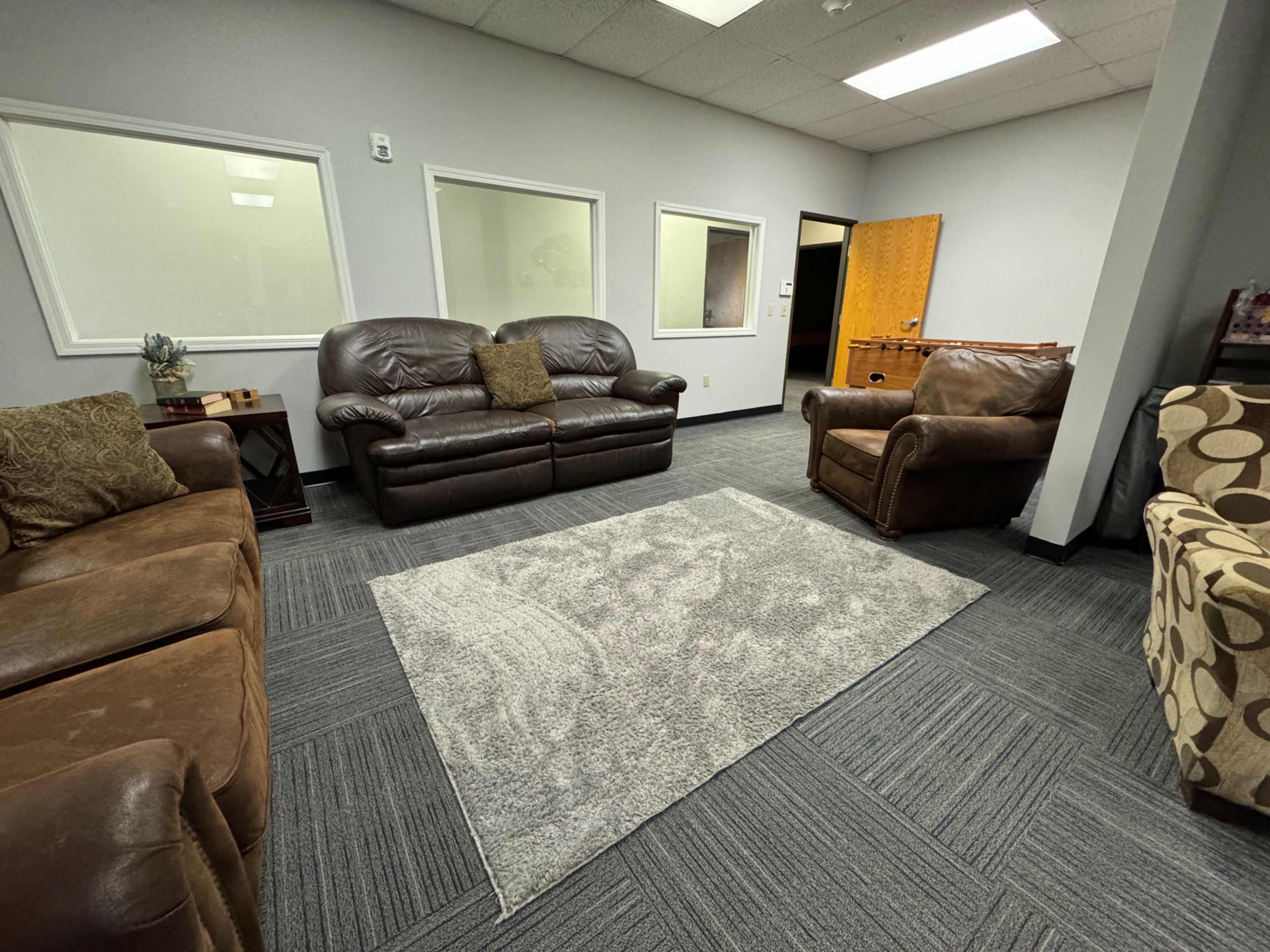 A waiting area with two brown leather sofas, a patterned chair, and a gray rug on a carpeted floor, enclosed by light-colored walls and a wooden door.