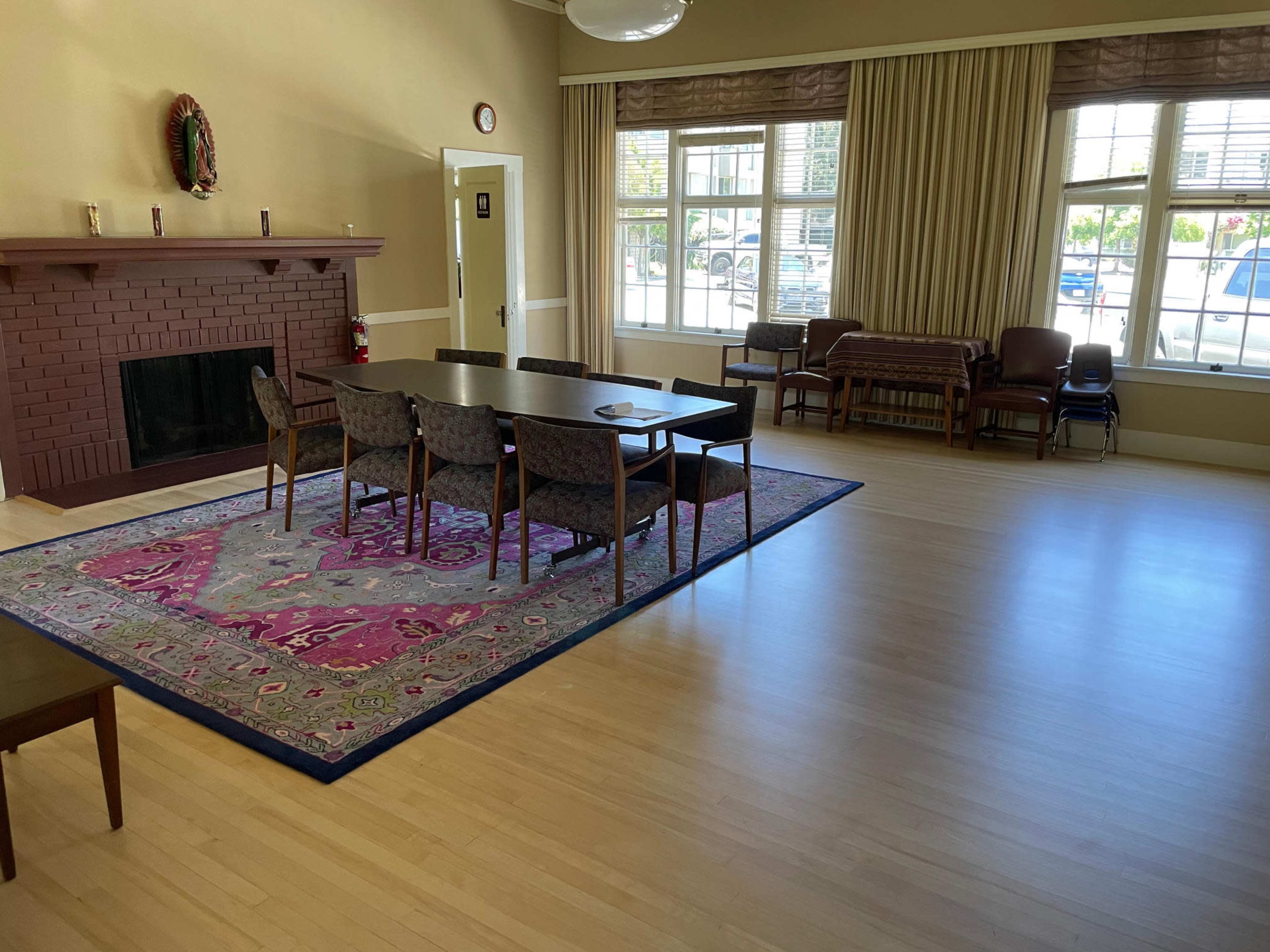 The room features a large table surrounded by chairs on a patterned rug, with a fireplace and windows letting in natural light.